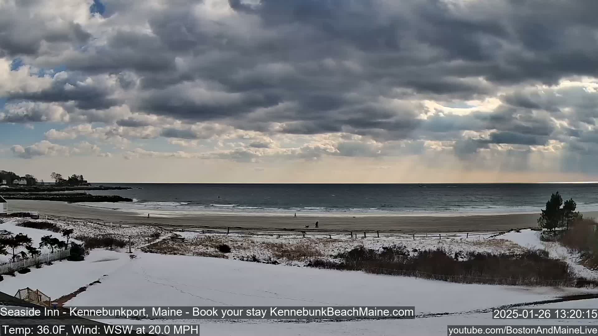 A snow-covered beach scene shows a partly cloudy sky with sunbeams breaking through, and people walking on the sandy shore near the ocean.