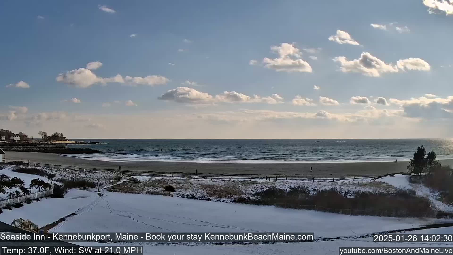 A snow-covered beach scene shows a partly cloudy sky, a calm ocean with small waves, and a few people walking on the sand.