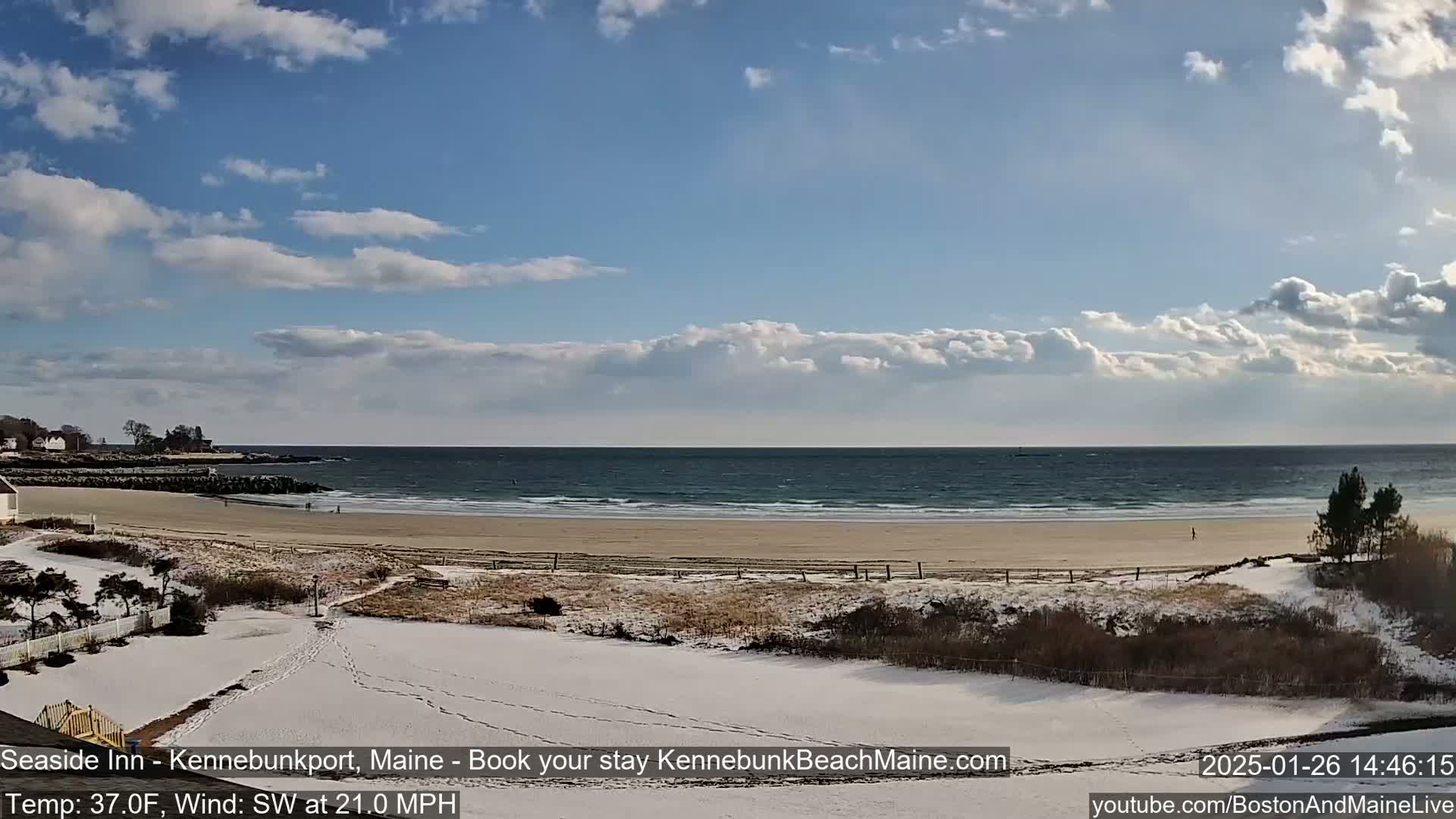 A partly cloudy day shows a sandy beach with a few people walking along the shore, partially covered in snow, with a calm ocean in the background.