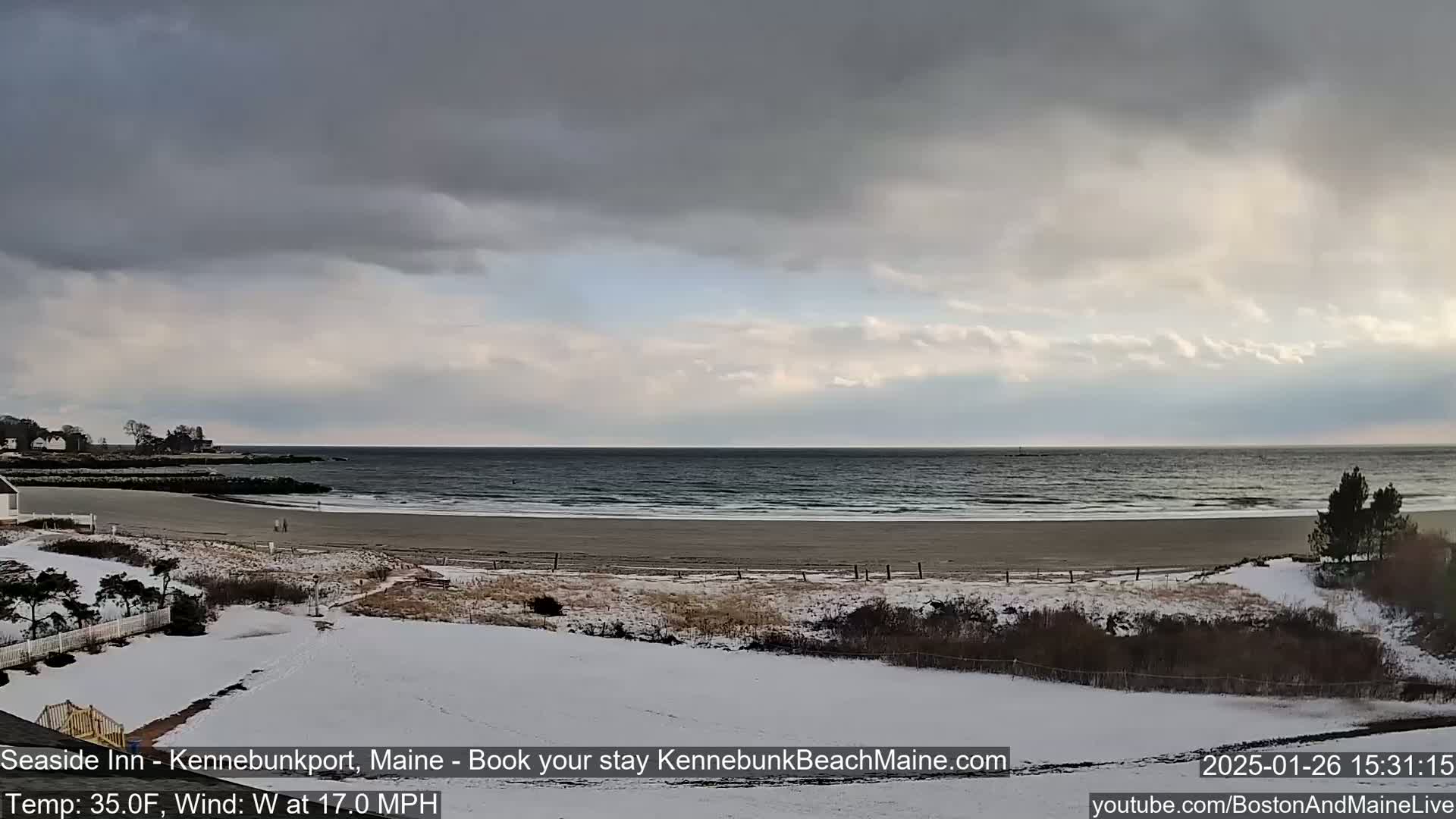 A snow-covered beach and coastline are visible under a mostly cloudy sky, with a moderate westerly wind.