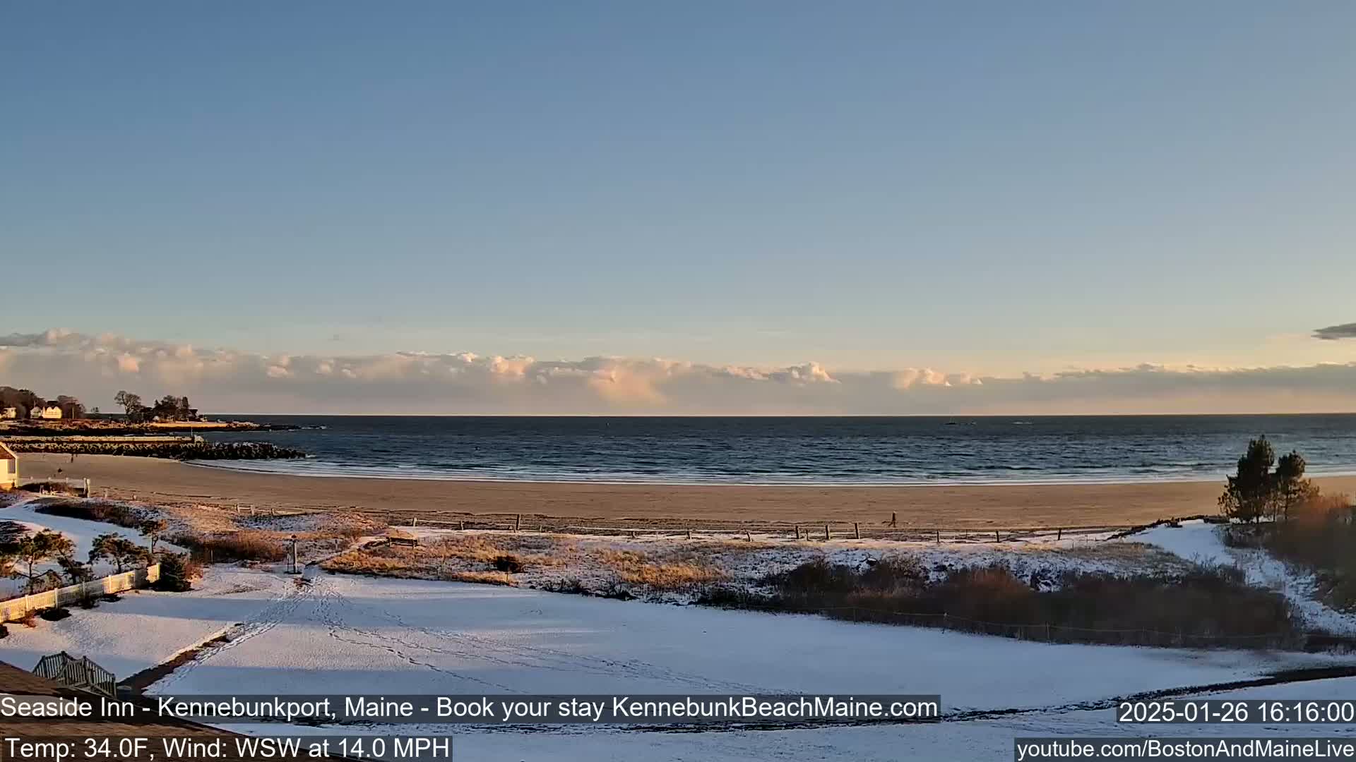 A snow-covered beach with a calm ocean under a partly cloudy sky is viewed from a slight elevation.
