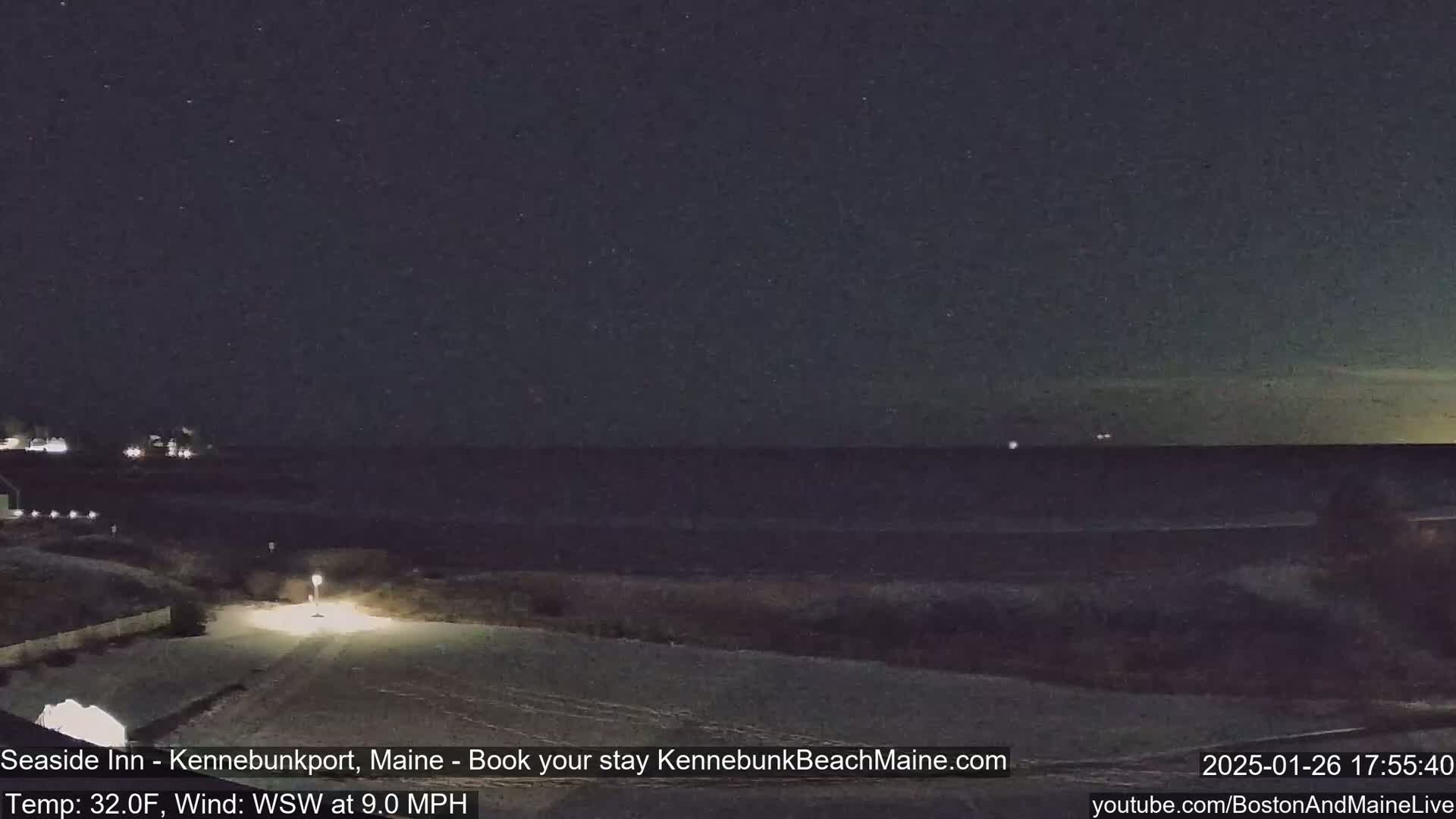 A nighttime view of a snow-covered coastal area shows a dark ocean under a starry sky, with a road and some lights visible in the foreground, and a 32°F temperature and 9 mph wind from the west-southwest.
