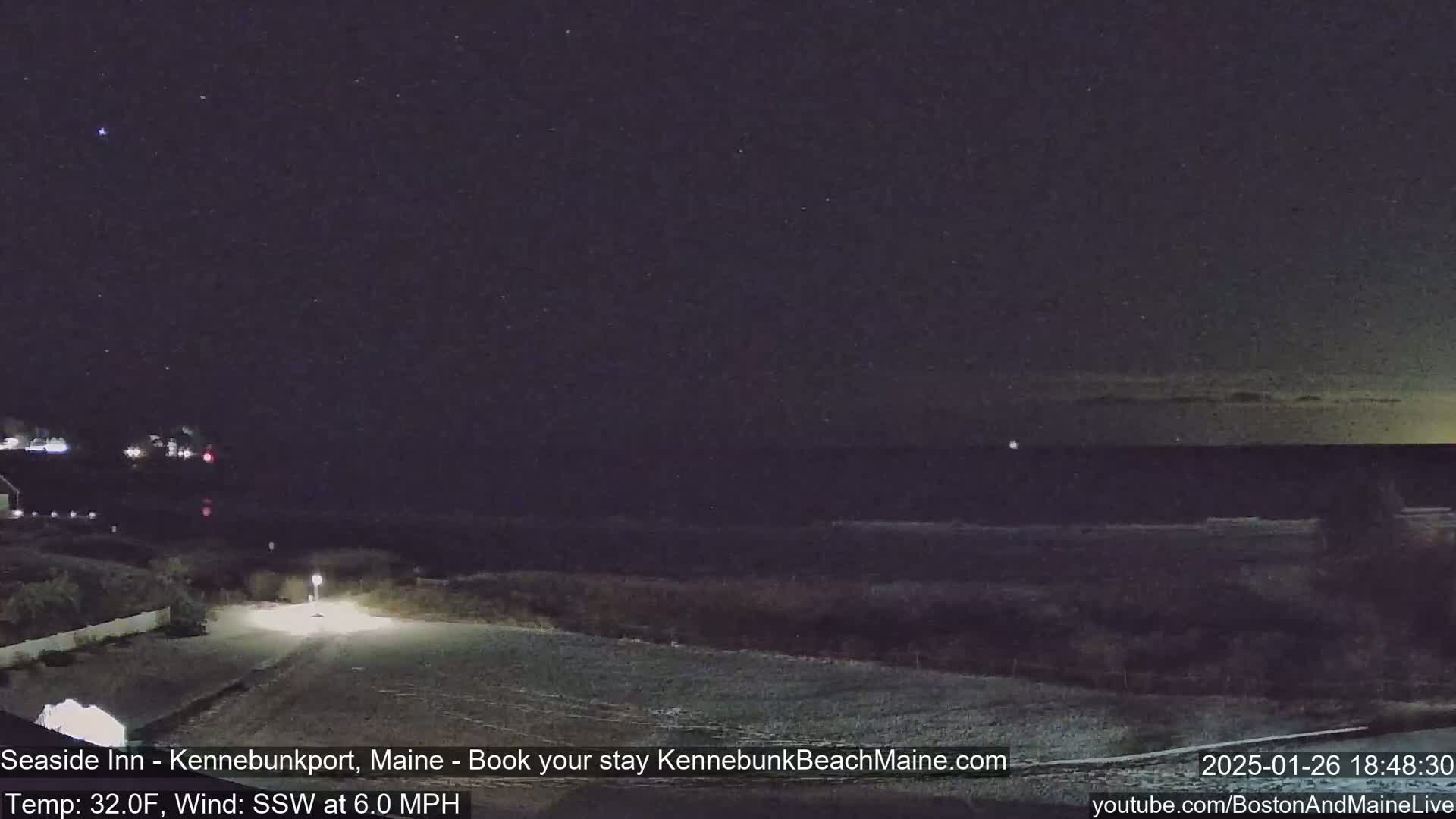 A nighttime coastal scene shows a dark ocean under a starry sky with a few clouds on the horizon, and a snow-covered roadway leading to the water's edge.