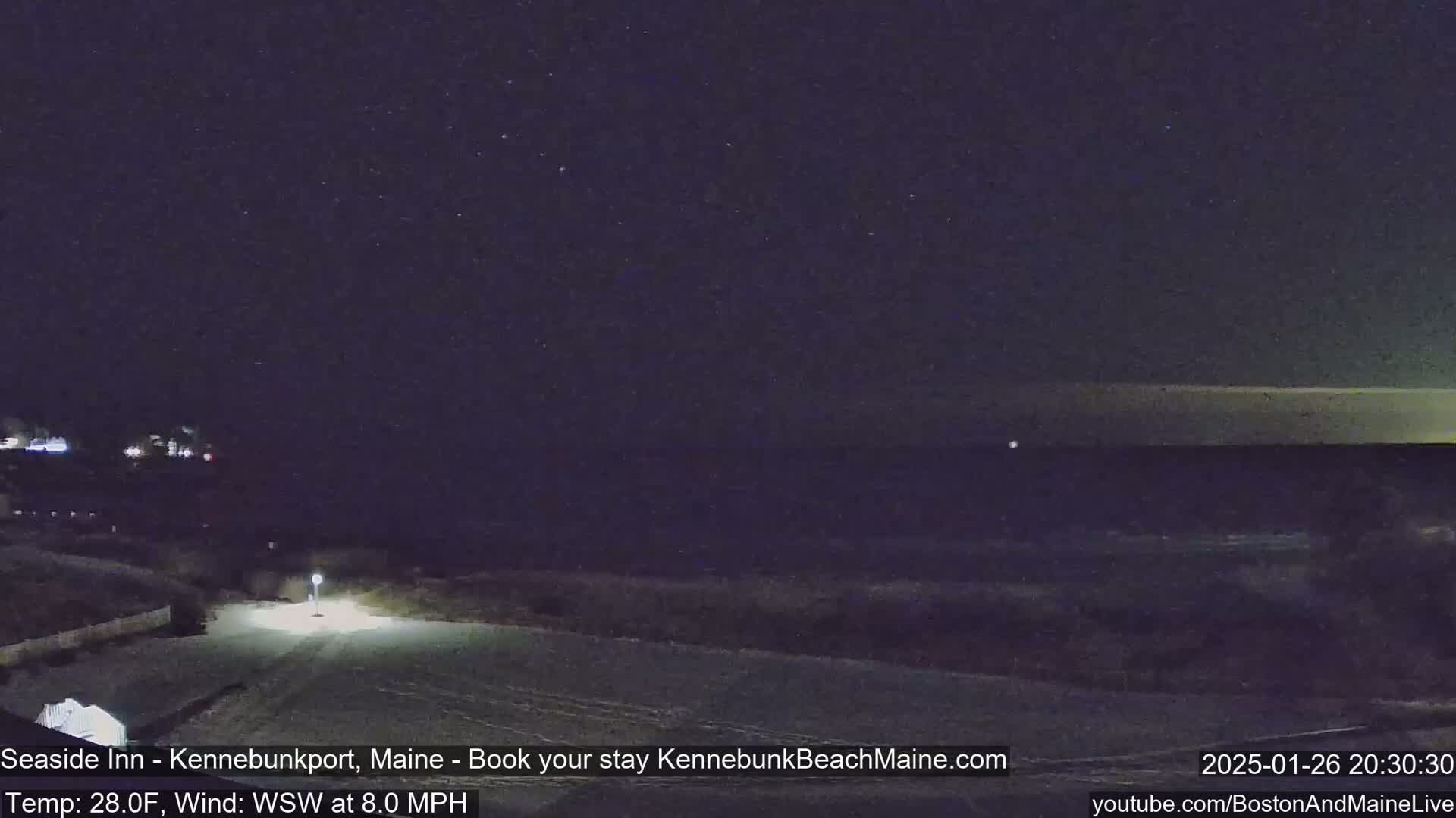 A nighttime view of a snow-covered road leading to a dark ocean under a starry sky, with a light wind.