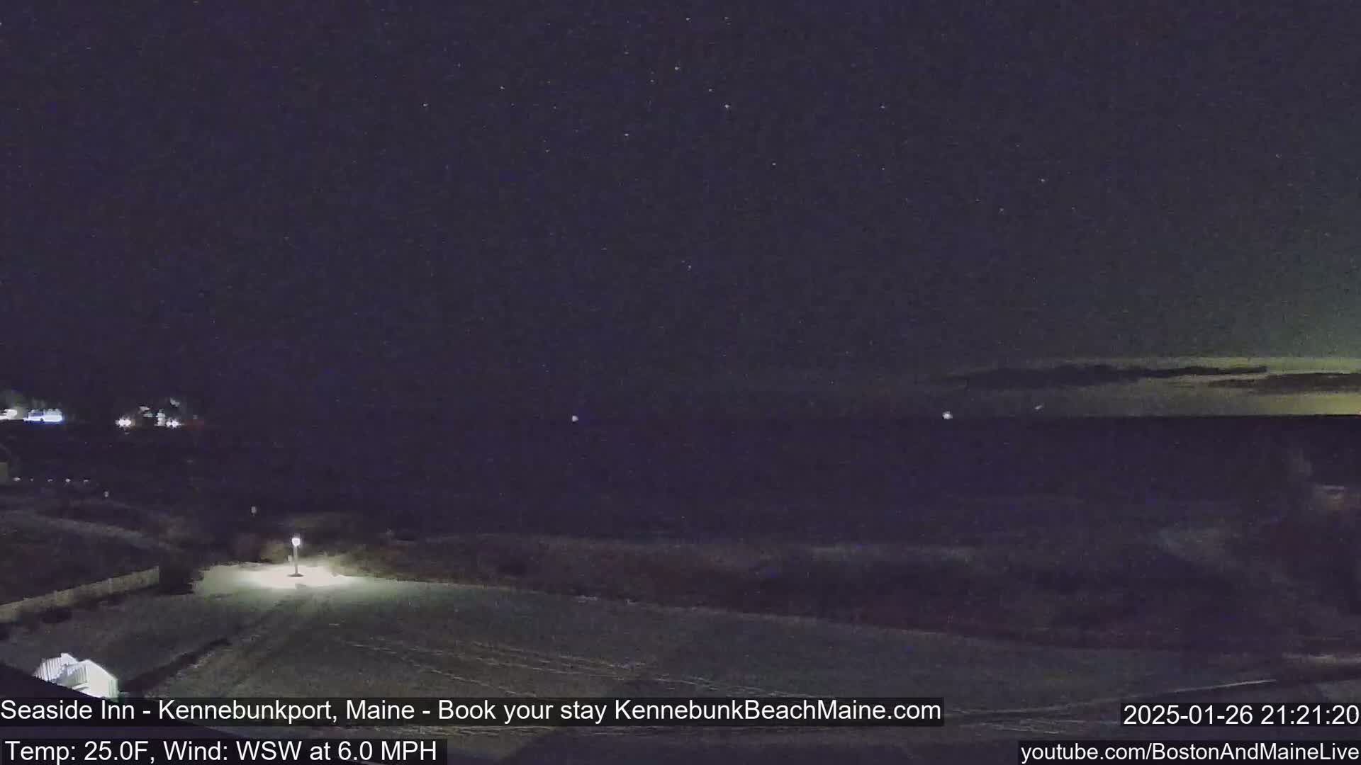 A nighttime view of a snow-covered coastal area under a partly cloudy sky shows a calm body of water in the distance.