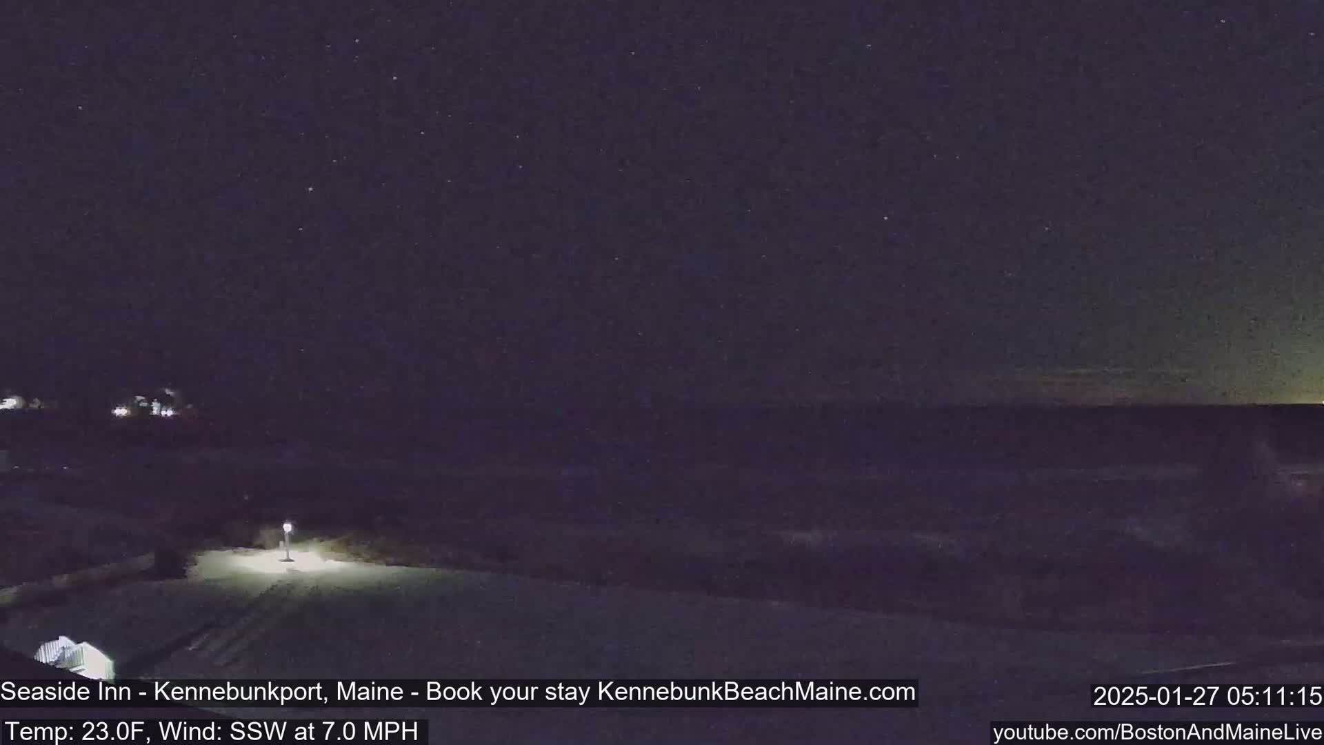 A nighttime, snow-covered scene shows a road leading to a dimly lit area under a starry sky with a southwest wind blowing at 7 mph and a temperature of 23 degrees Fahrenheit.
