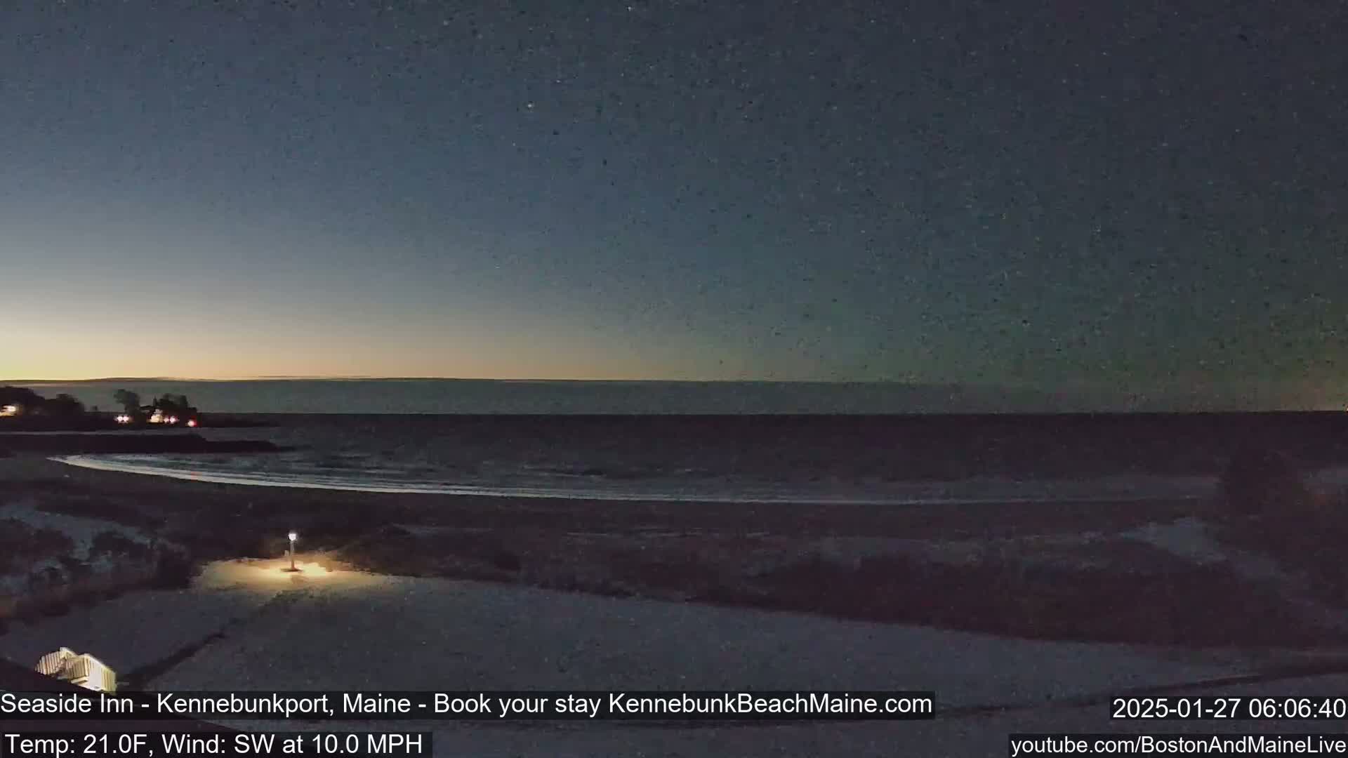 A dark, snowy beach is shown at night with a calm ocean under a twilight sky, with a southwest wind blowing at 10 mph and a temperature of 21 degrees Fahrenheit.