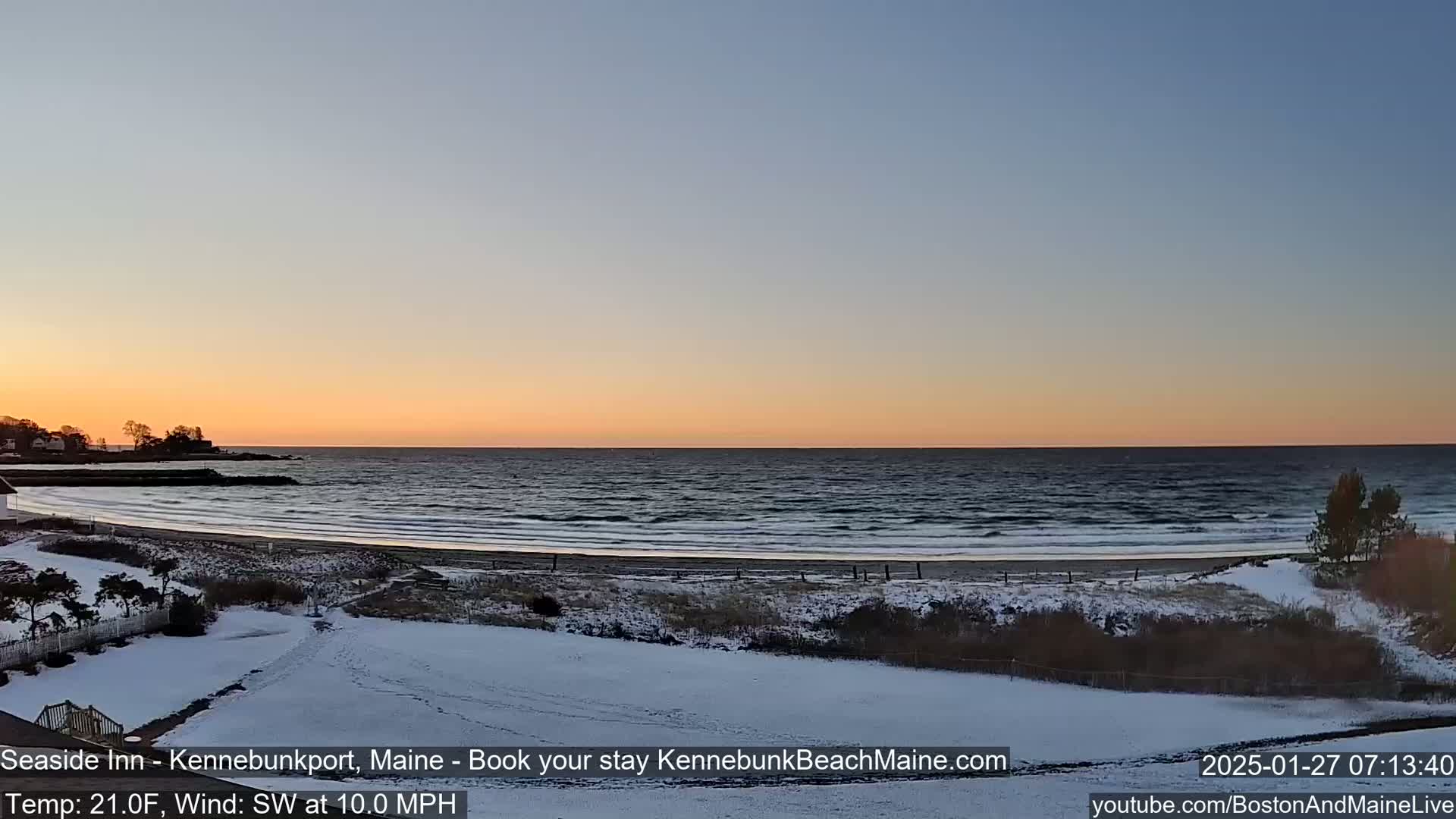 A snow-covered beach scene at sunset shows a calm ocean with small waves under a clear, light-orange sky.