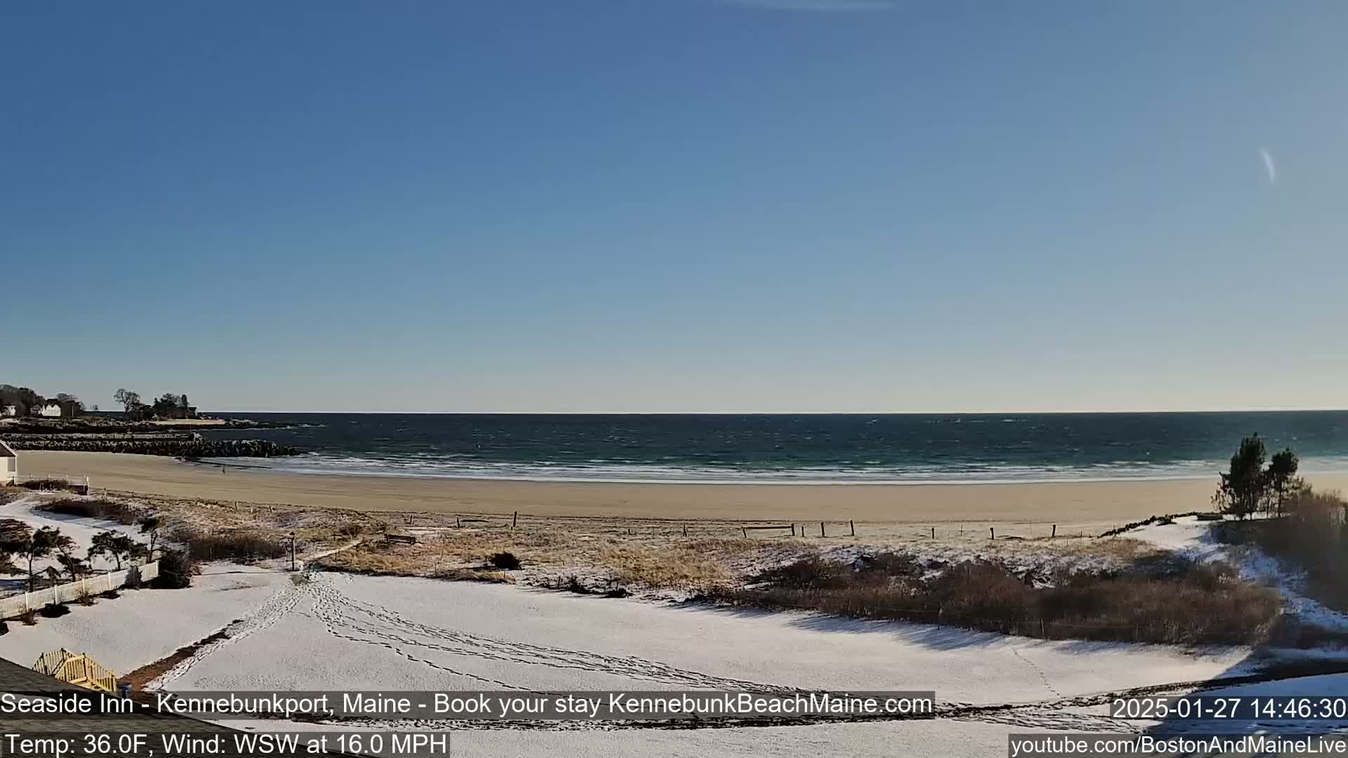 A sandy beach with a partly snow-covered foreground and a wavy ocean under a clear blue sky.