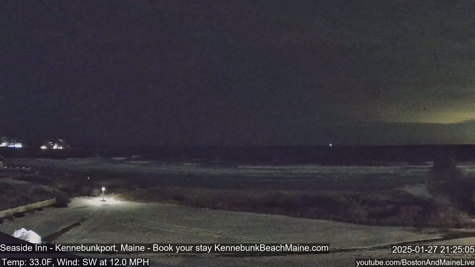 A snow-covered coastal area at night shows a dark ocean with some waves under a mostly clear, dark sky.