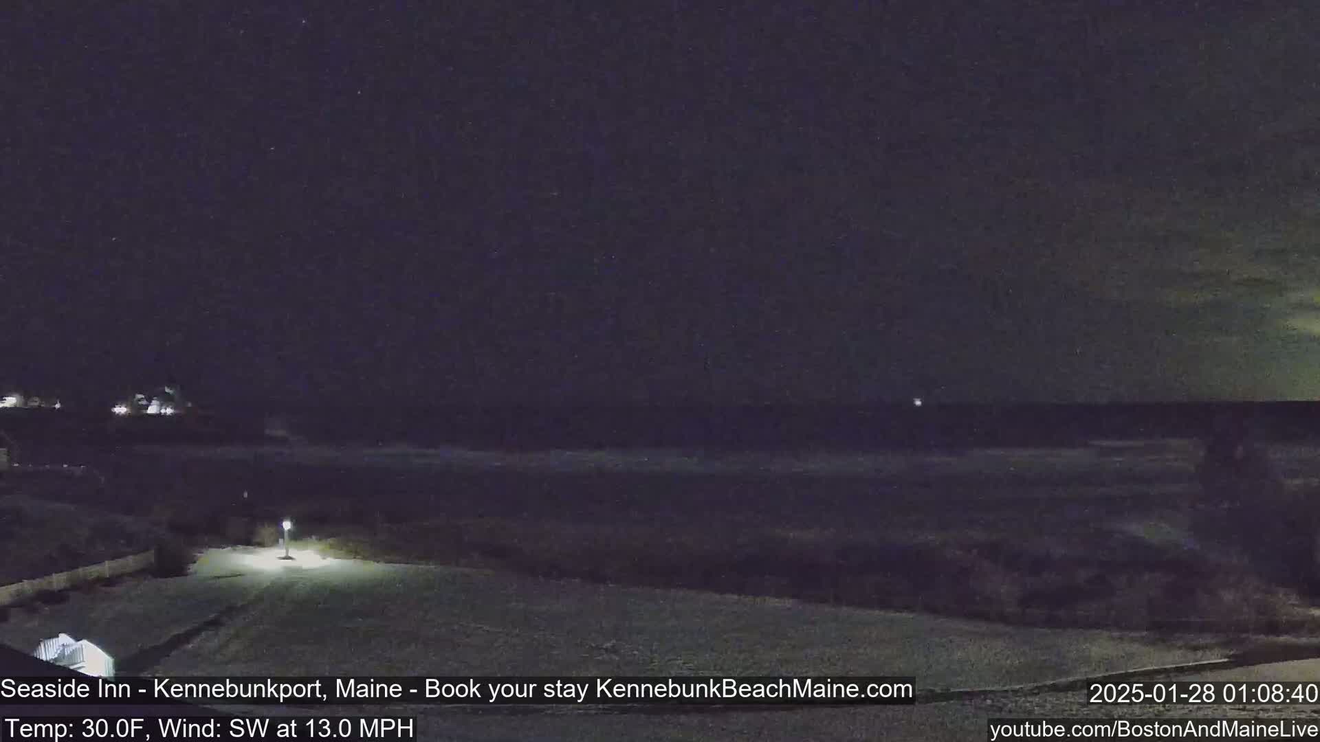 A snow-covered landscape at night shows a dark ocean and a dimly lit path leading to a building under a dark sky with a southwest wind of 13 mph and a temperature of 30°F.