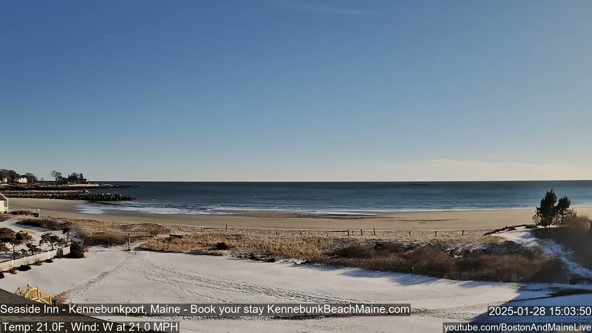 A sandy beach partially covered in snow stretches along a calm ocean under a clear blue sky, with a light dusting of snow on the ground in the foreground.