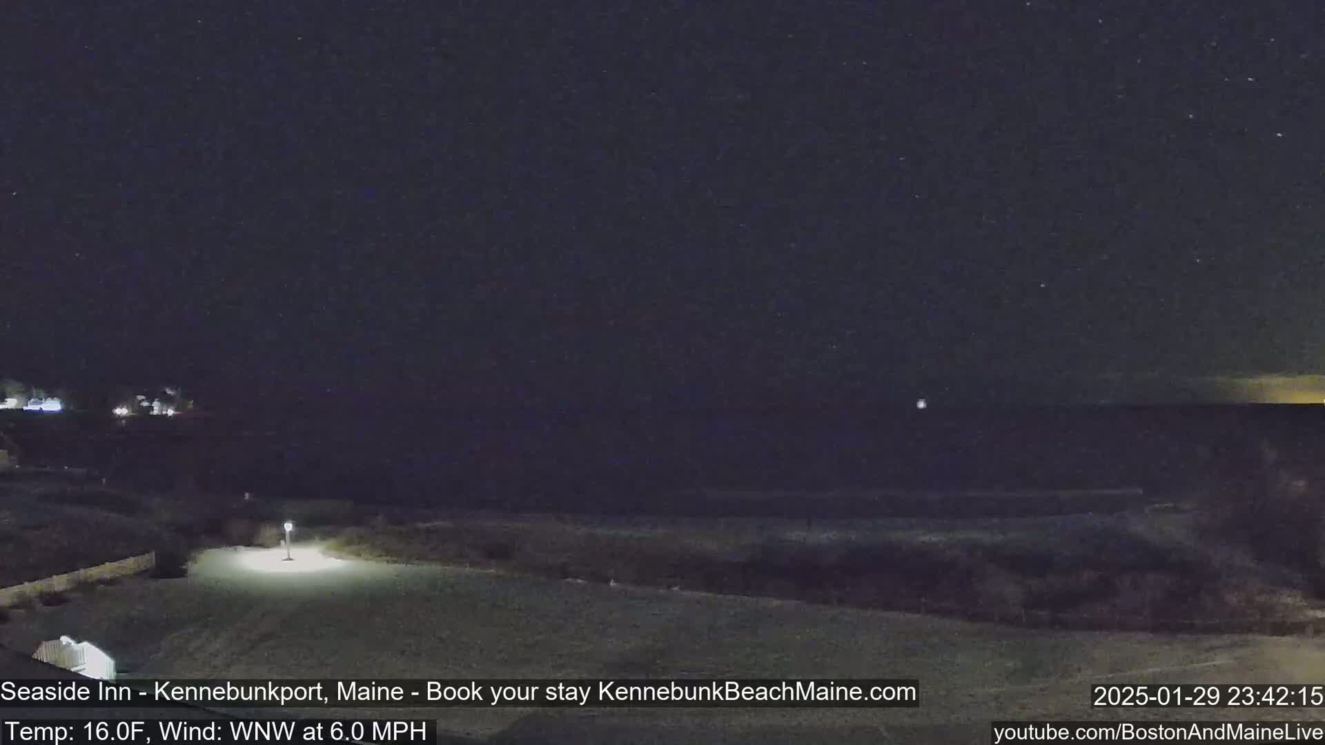 A nighttime view of a beach with a starry sky, a calm ocean, and a single lamp illuminating a paved area.