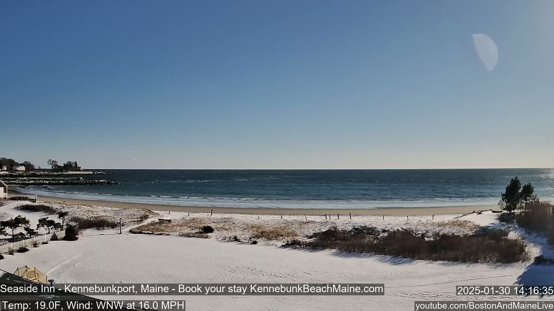 A snow-covered beach with a partially visible crescent moon in a clear blue sky, overlooking a wavy ocean with a brisk wind.