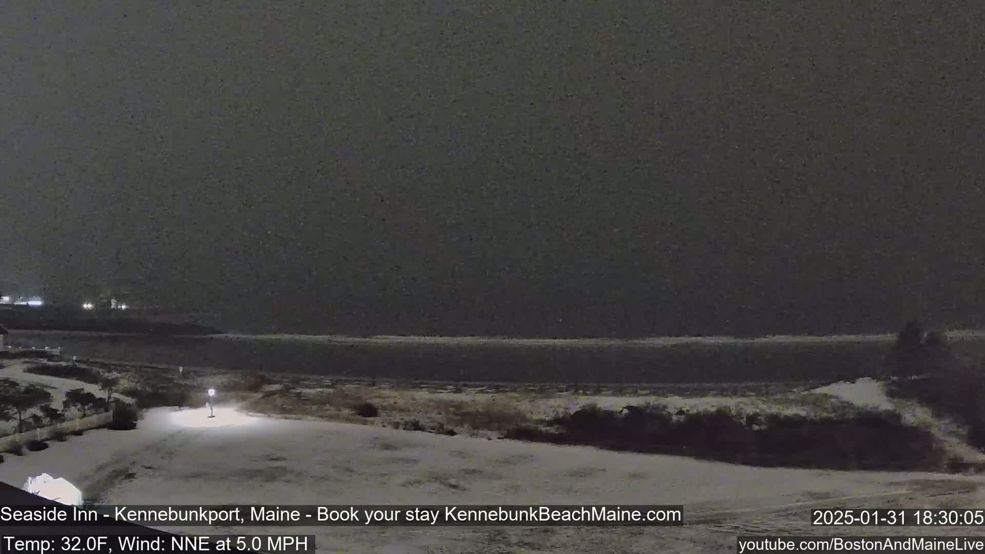 A snow-covered landscape at night shows a dark body of water under a gray sky, with a single lit lamppost in the foreground.