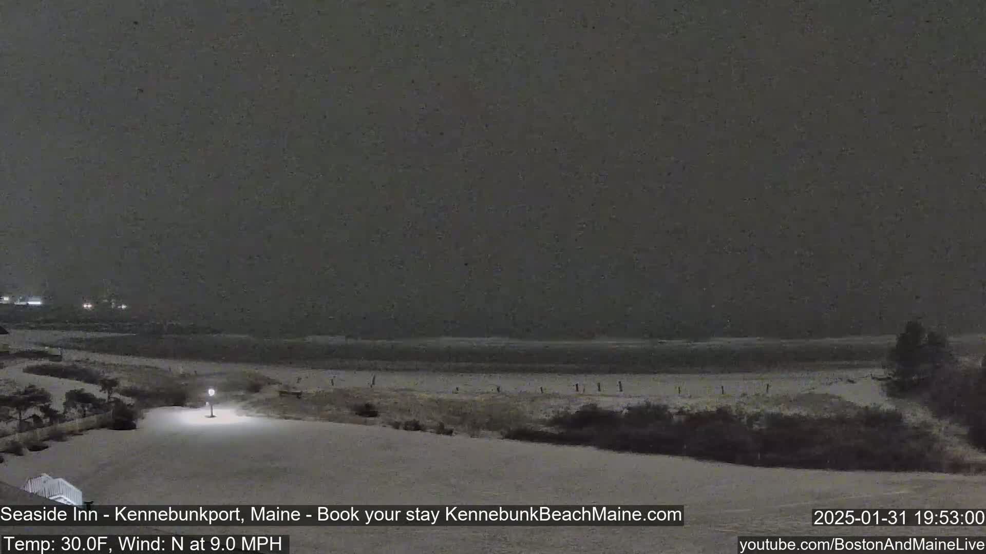 A snow-covered beach at night with a dark, overcast sky and a faint light from a lamppost.