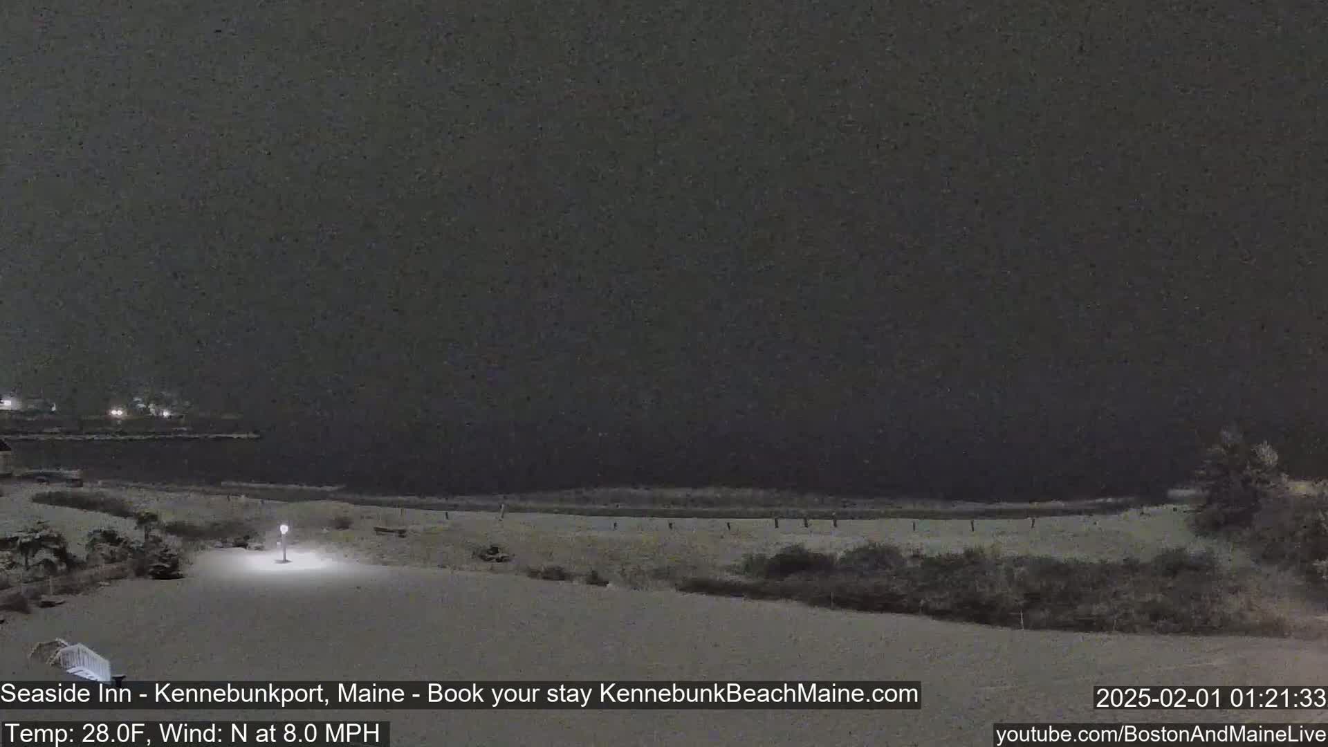 A snow-covered beach at night is illuminated by a single lamppost, with a dark body of water visible in the background under a dark sky.