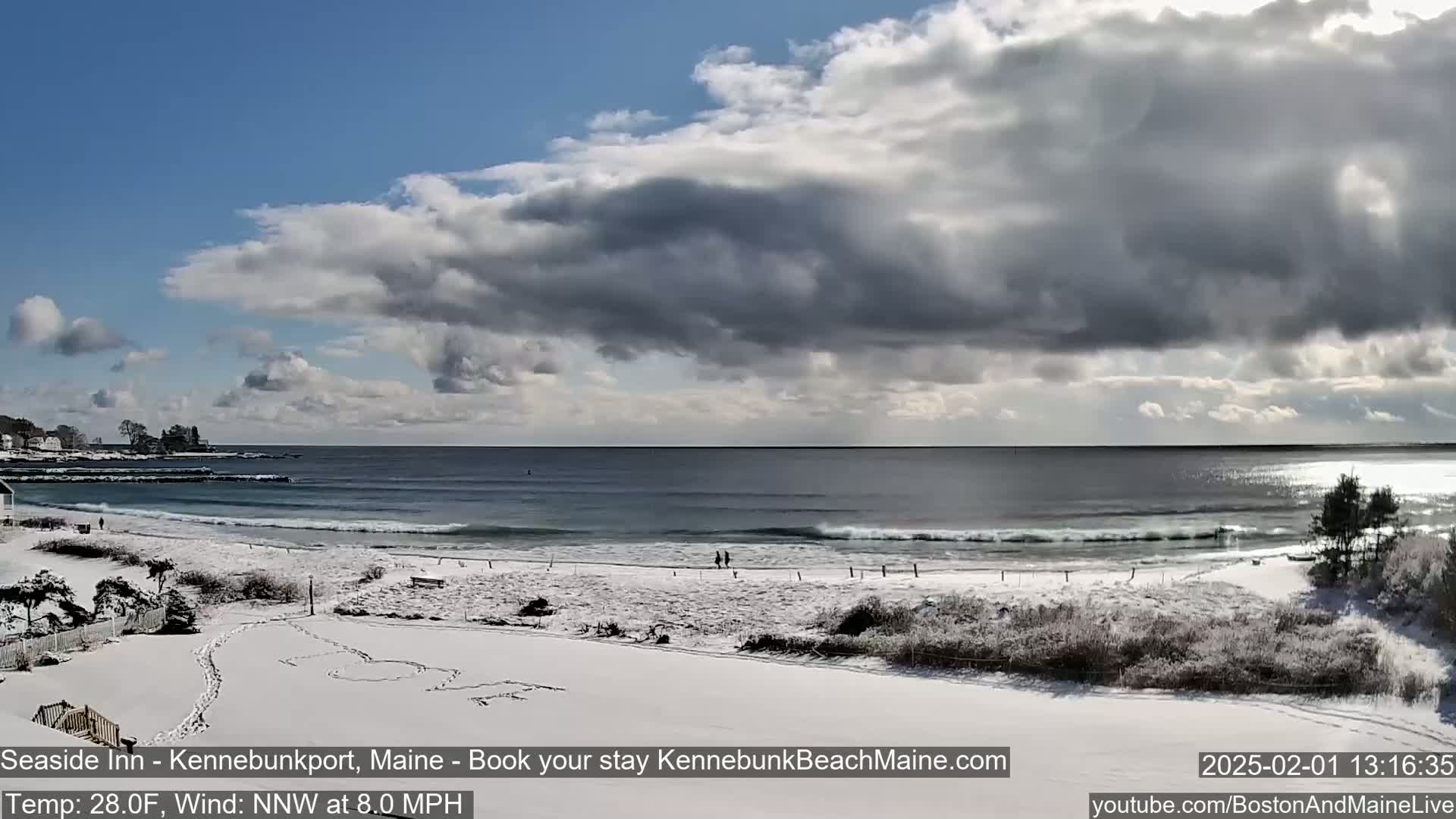 A snow-covered beach with a partly cloudy sky and a calm ocean shows small waves breaking on the shore.