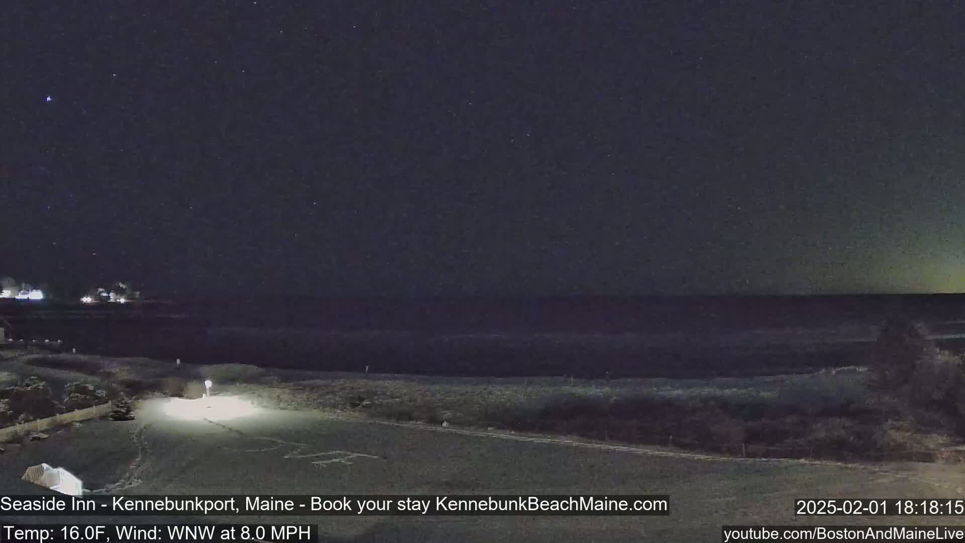 A nighttime, coastal scene shows a dark ocean under a starry sky, with a snow-covered road and some distant lights in the foreground, and an 8 mph wind.