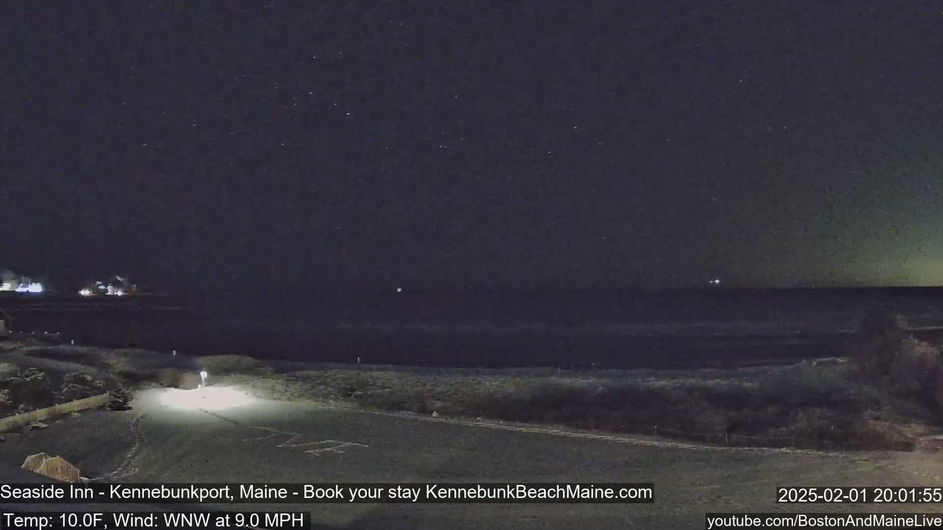 A nighttime view of a dark ocean under a starry sky, with a sparsely lit road leading to the water's edge and a 10°F temperature with a 9 mph wind from the west-northwest.