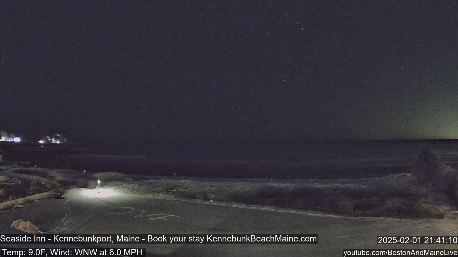 A nighttime coastal scene shows a dark ocean under a starry sky, with a dimly lit paved area and grassy vegetation in the foreground.
