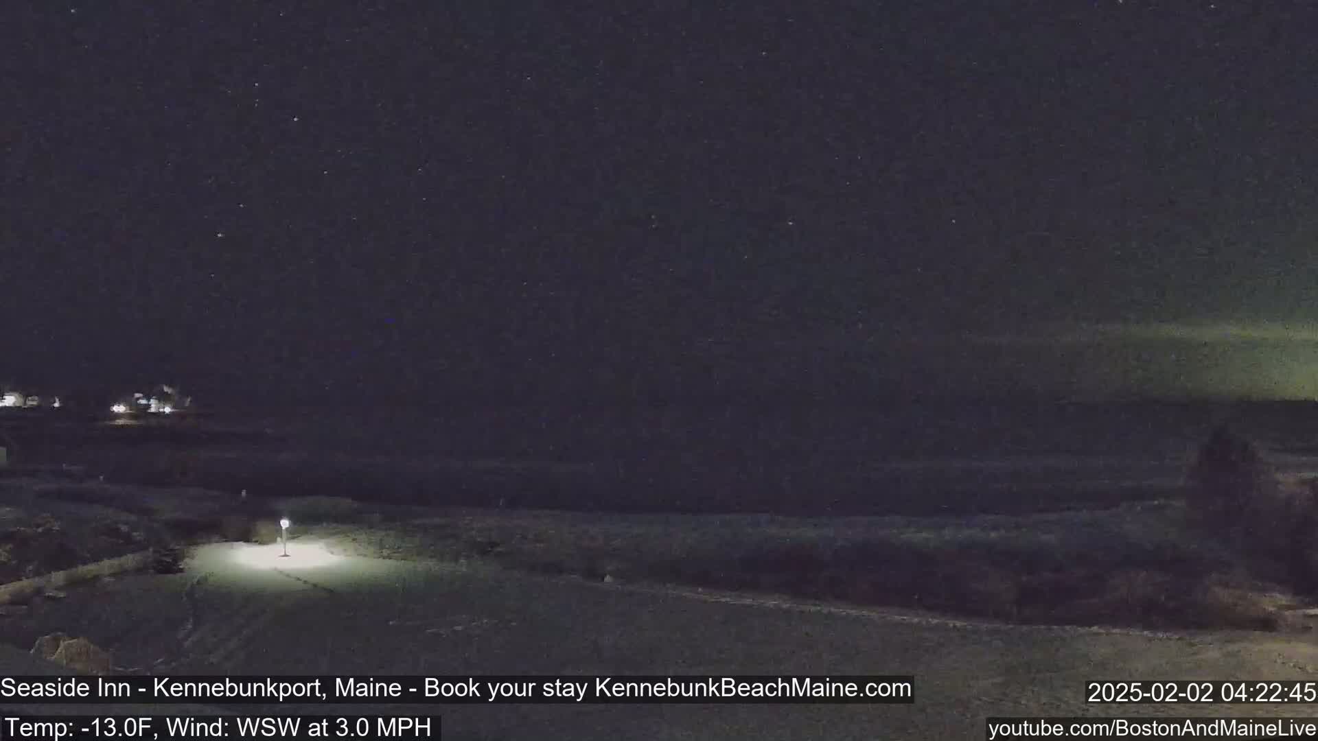 A nighttime, snow-covered beach scene under a starry sky with a faint aurora borealis, lit by a single lamppost.