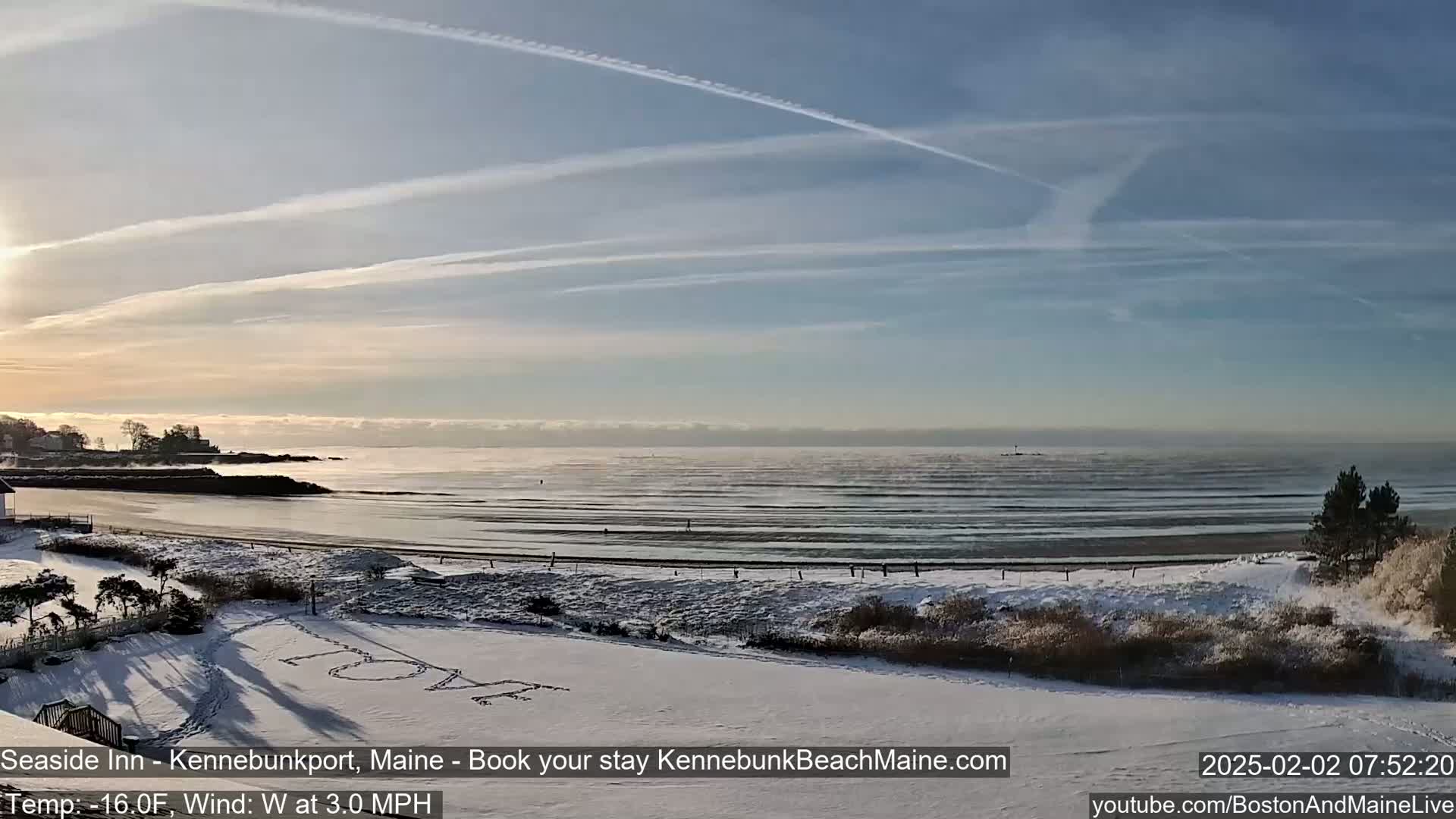 A snow-covered beach with the word "LOVE" written in the snow is seen, overlooking a calm ocean under a clear sky with contrails.