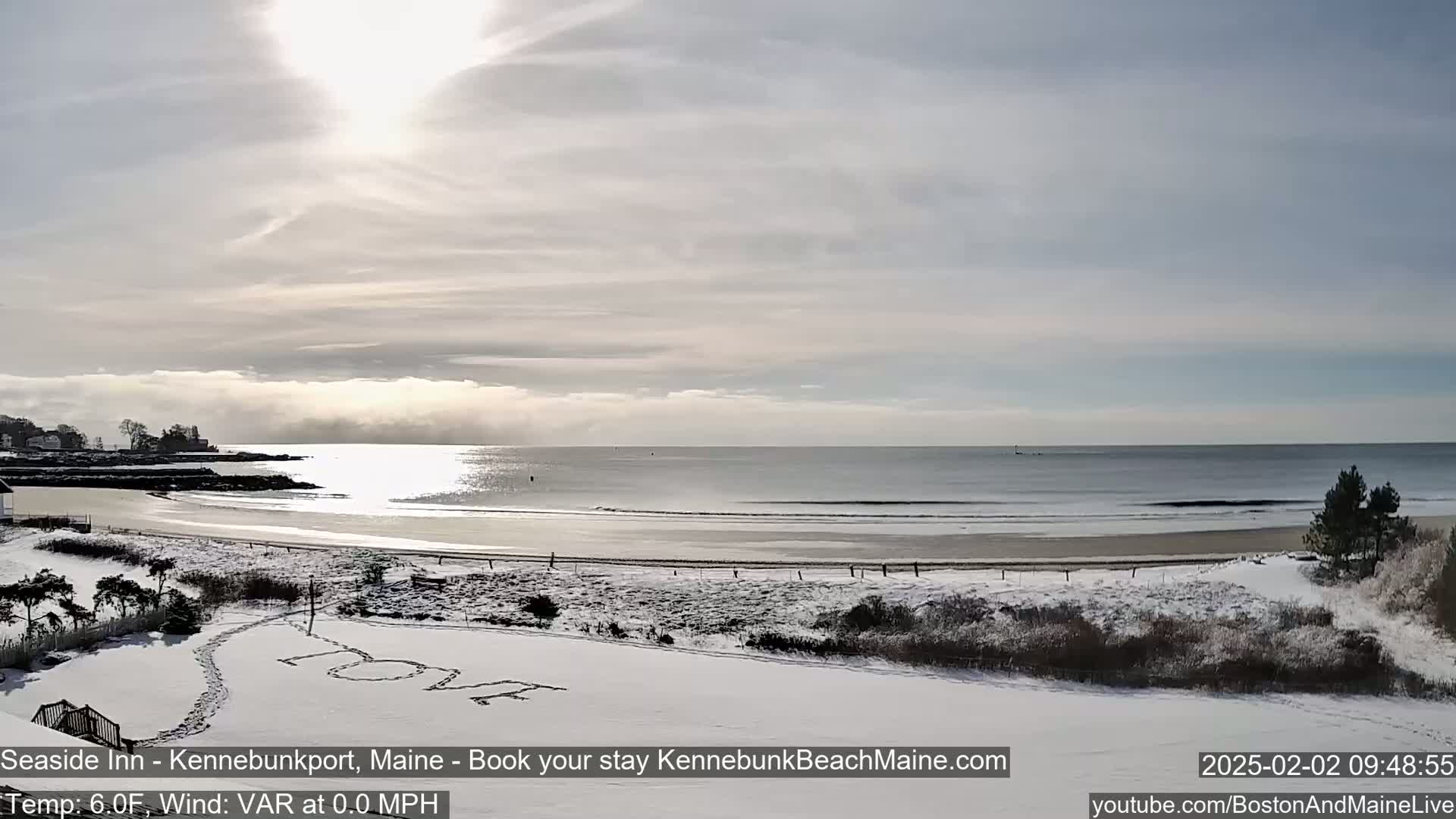 A snow-covered beach with calm waves and a partly cloudy sky is viewed from a snowy vantage point, showing the word "LOVE" written in the snow.