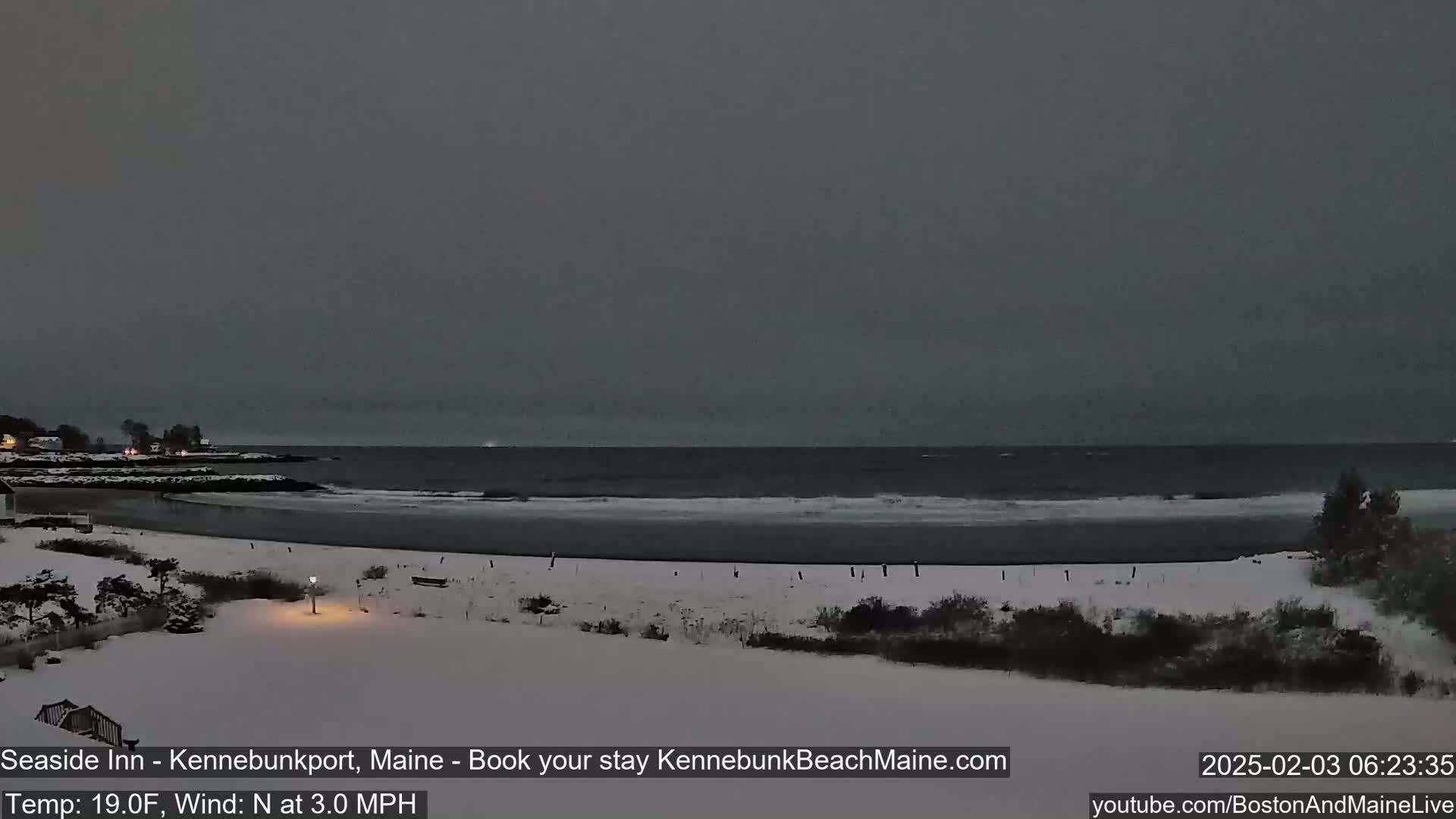 A snow-covered beach and shoreline are visible under a dark, overcast sky, with a calm ocean in the background.