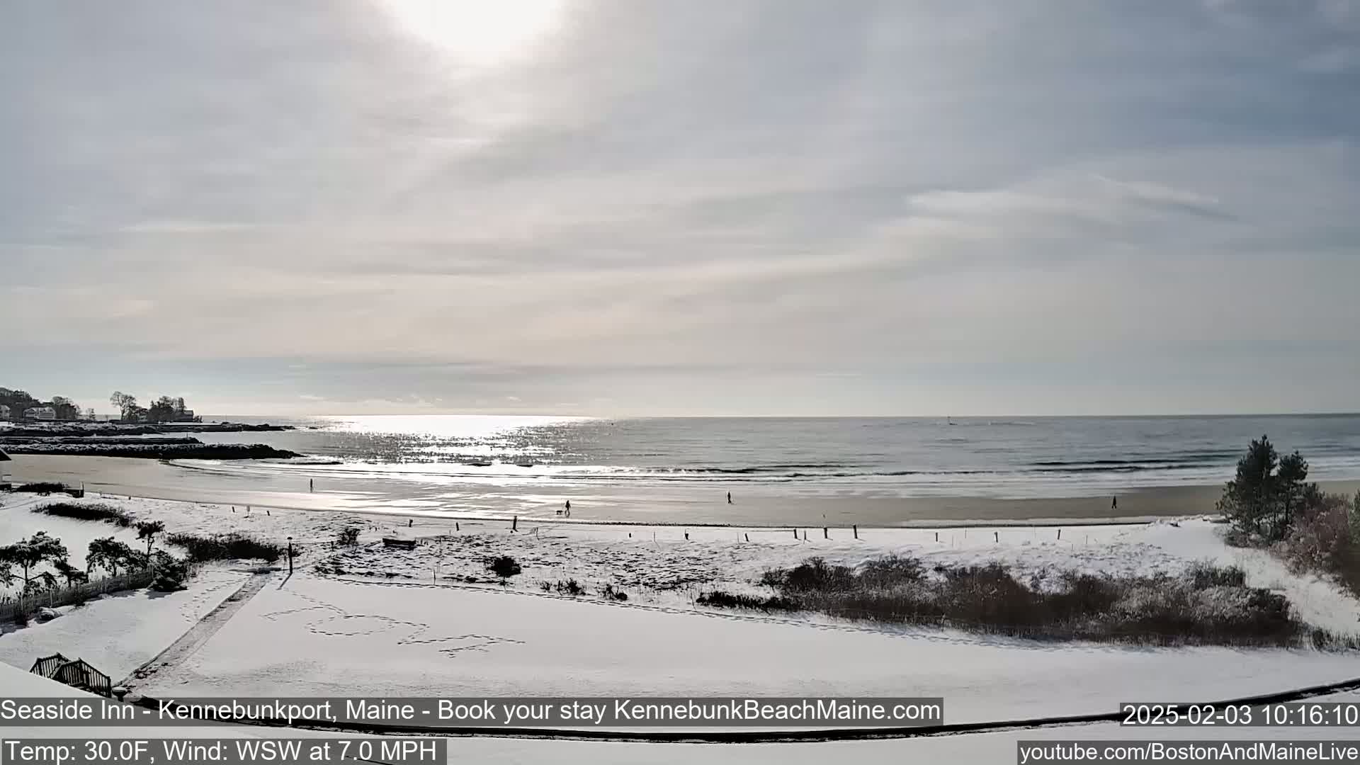 A snow-covered beach with a calm ocean under a bright, partly cloudy sky.