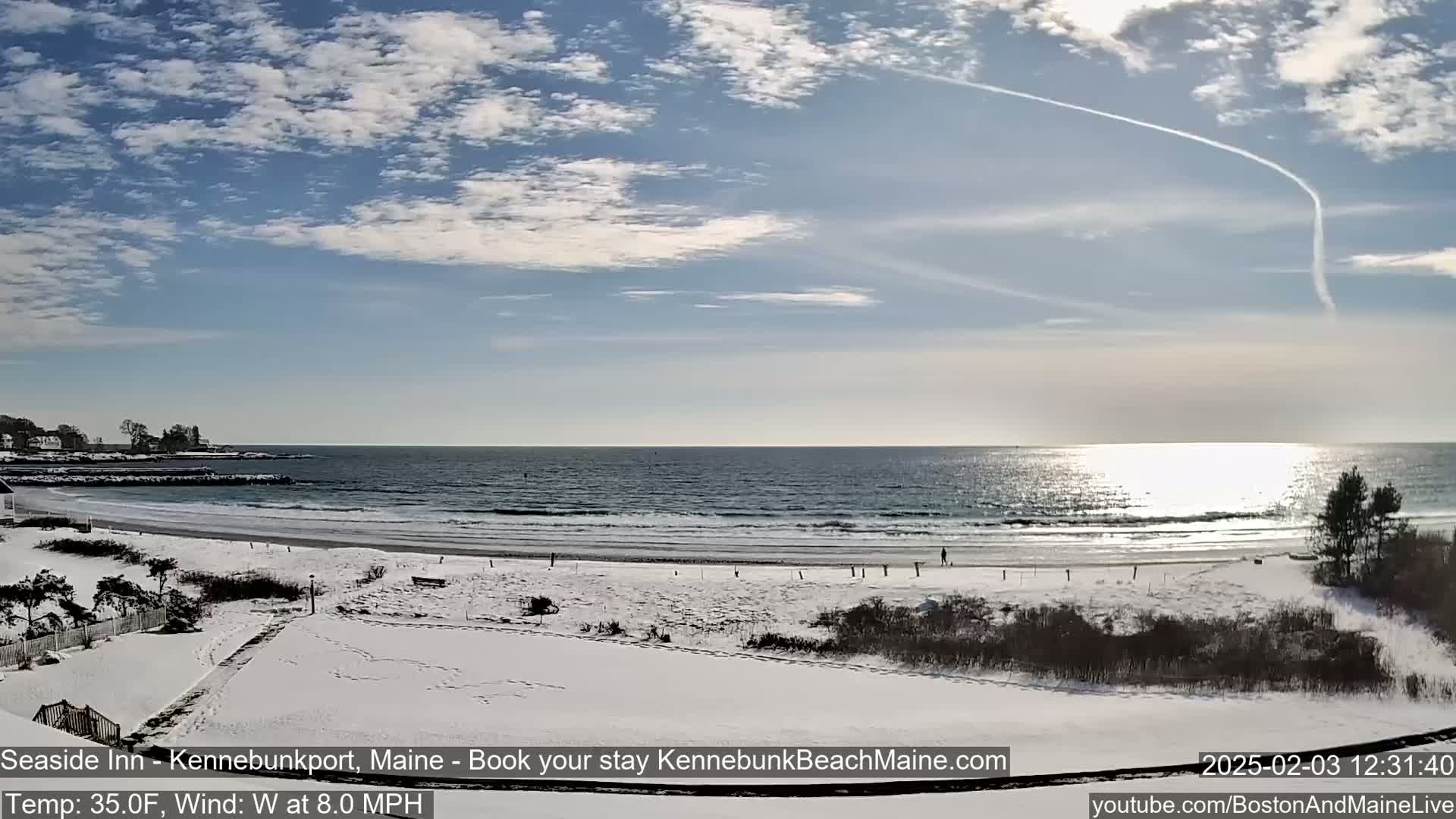 A snow-covered beach with a calm ocean and a partly cloudy sky under bright sunlight.