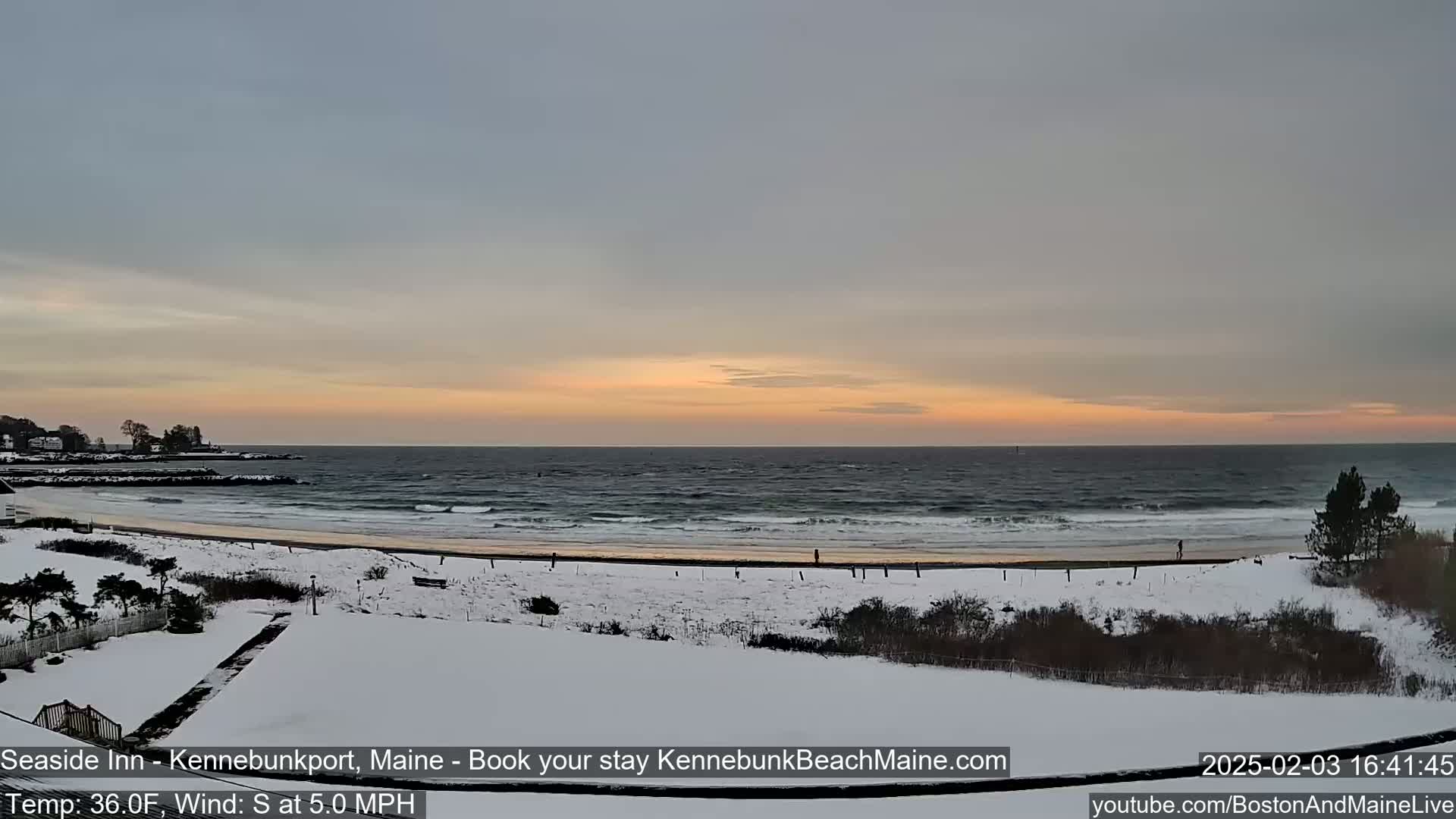 A snow-covered beach with a calm ocean under a pale orange sunset.