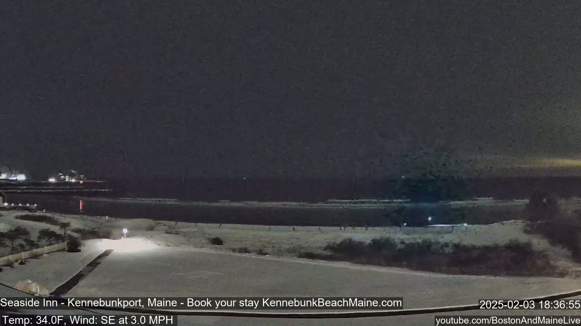 A nighttime coastal scene shows a dark ocean with small waves, a snow-covered beach with a few people, and a parking lot under a dark sky.