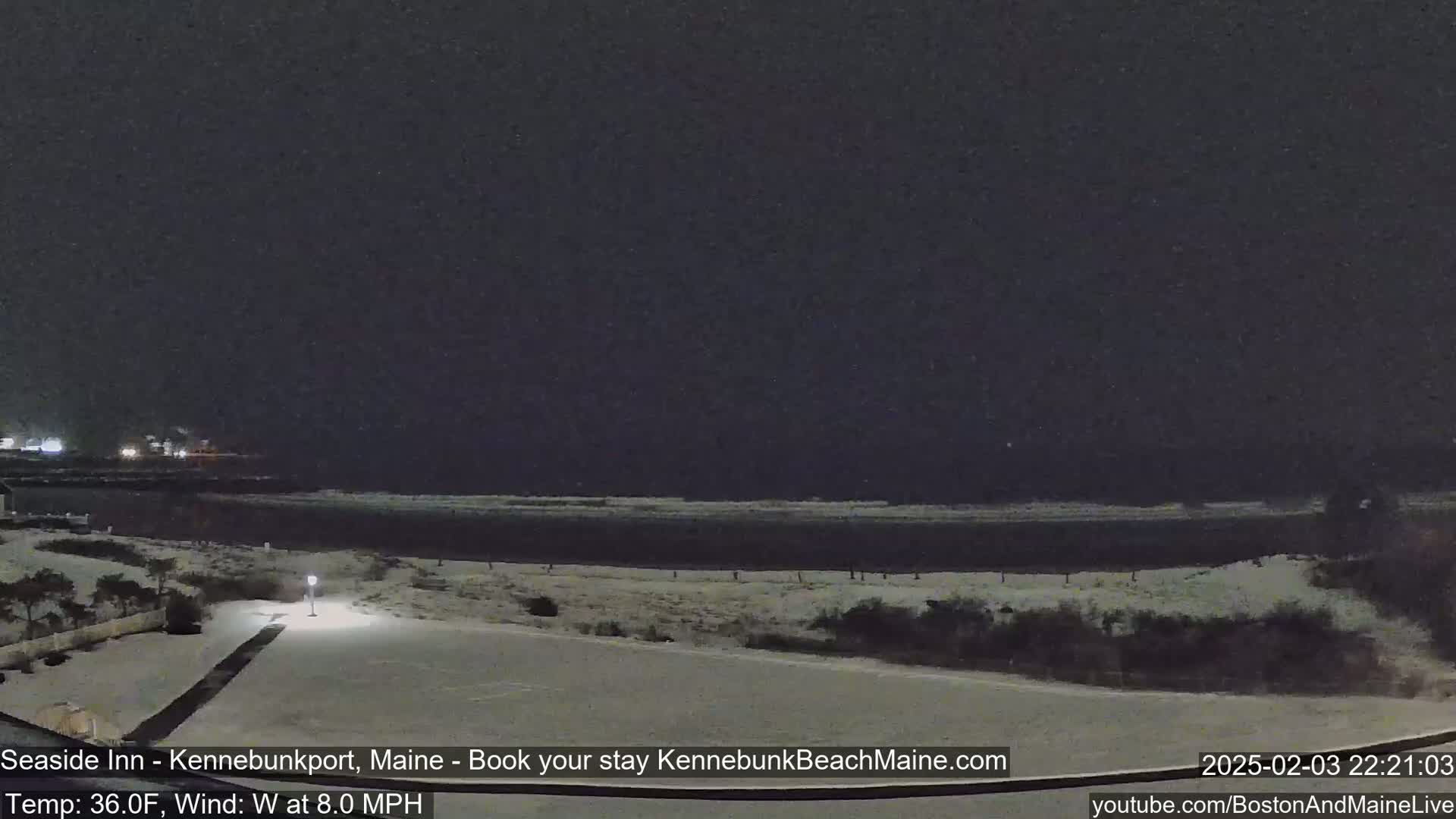 A snow-covered coastal scene at night is shown, with a calm body of water under a dark sky and a faint light from a lamppost.