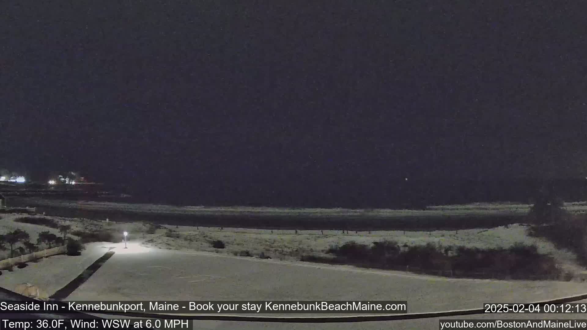 A snow-covered coastal scene at night, with a dark ocean and faint lights in the distance, under a dark sky.