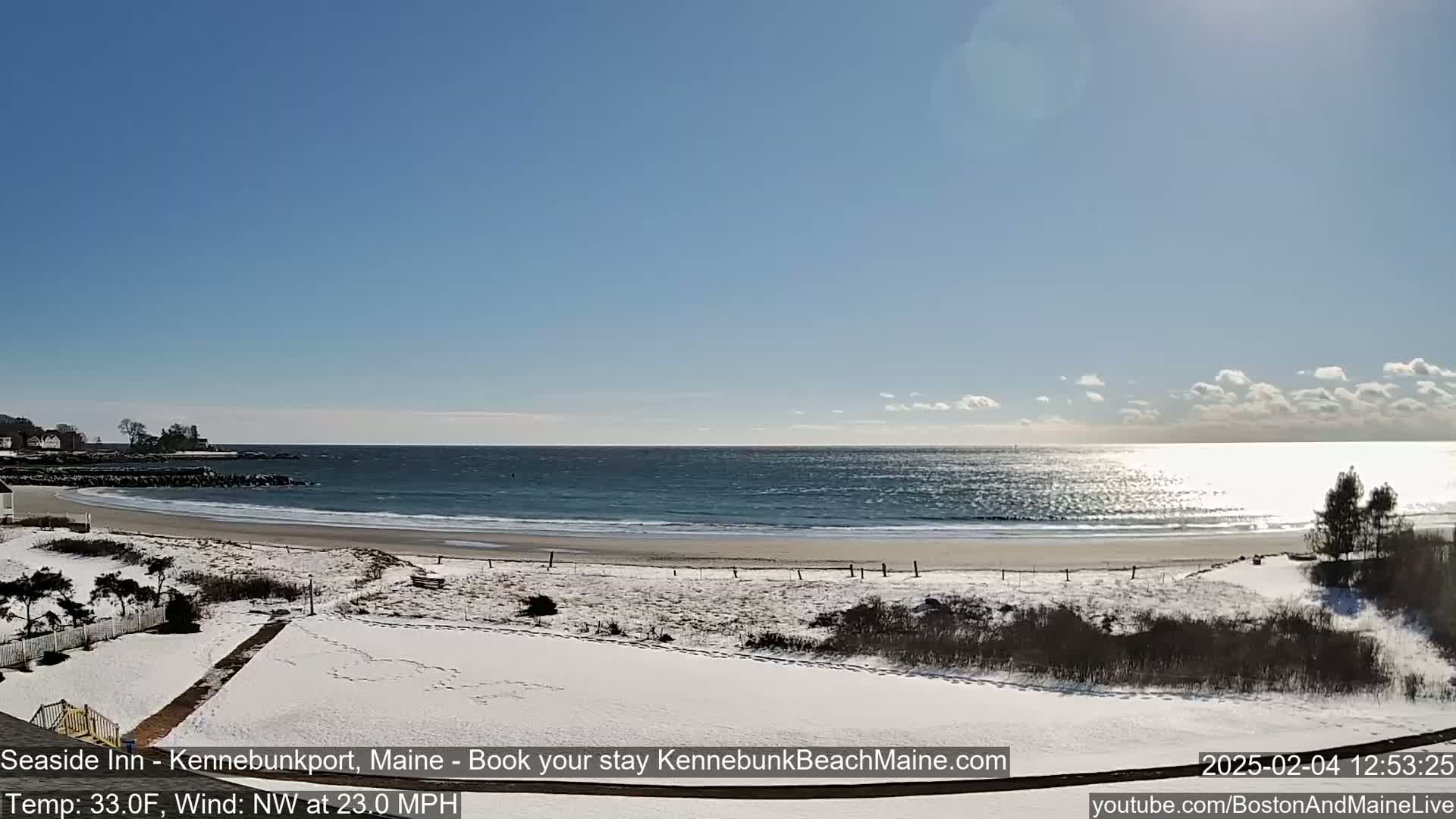 A snow-covered beach with a calm ocean under a mostly sunny sky is viewed from a snowy vantage point.