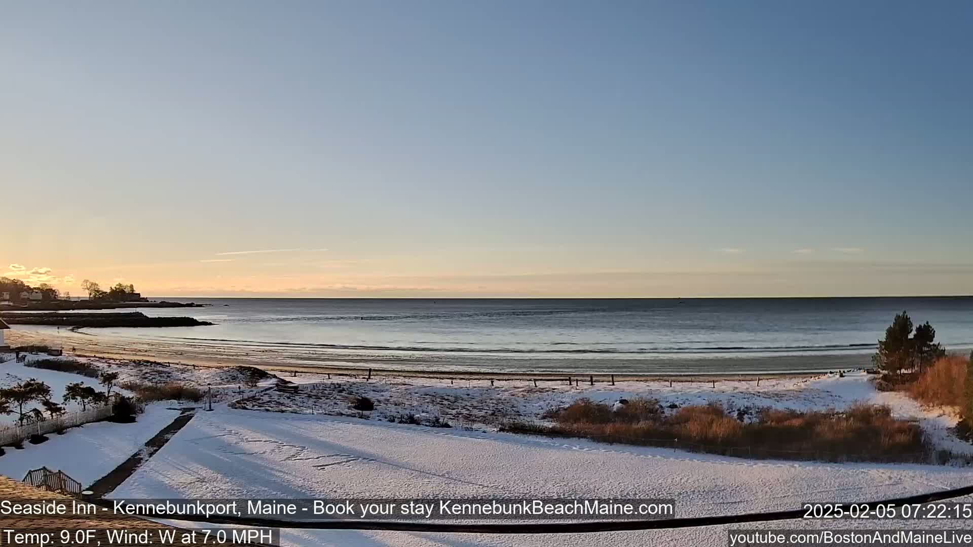 A snow-covered beach and shoreline are visible at sunrise, with calm ocean waves and a clear sky.