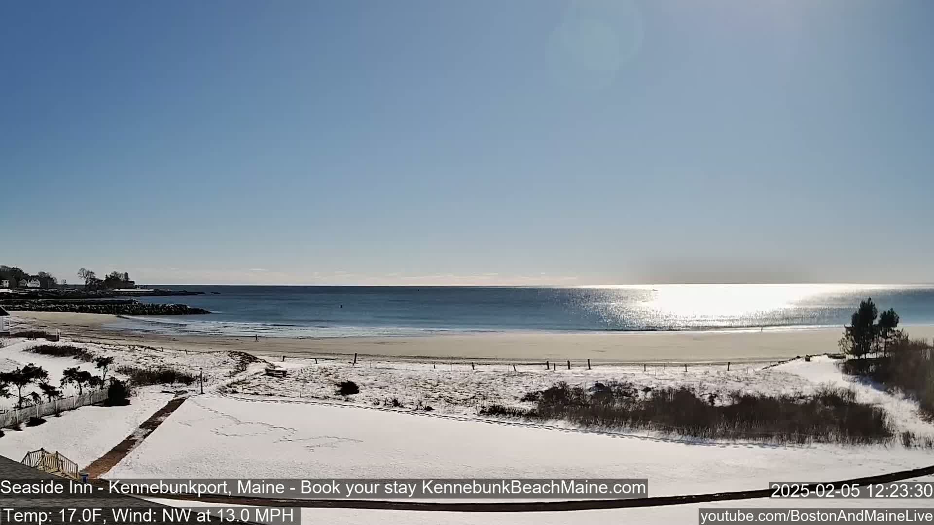 A snow-covered beach and grassy area are seen overlooking a calm ocean under a clear, sunny sky.