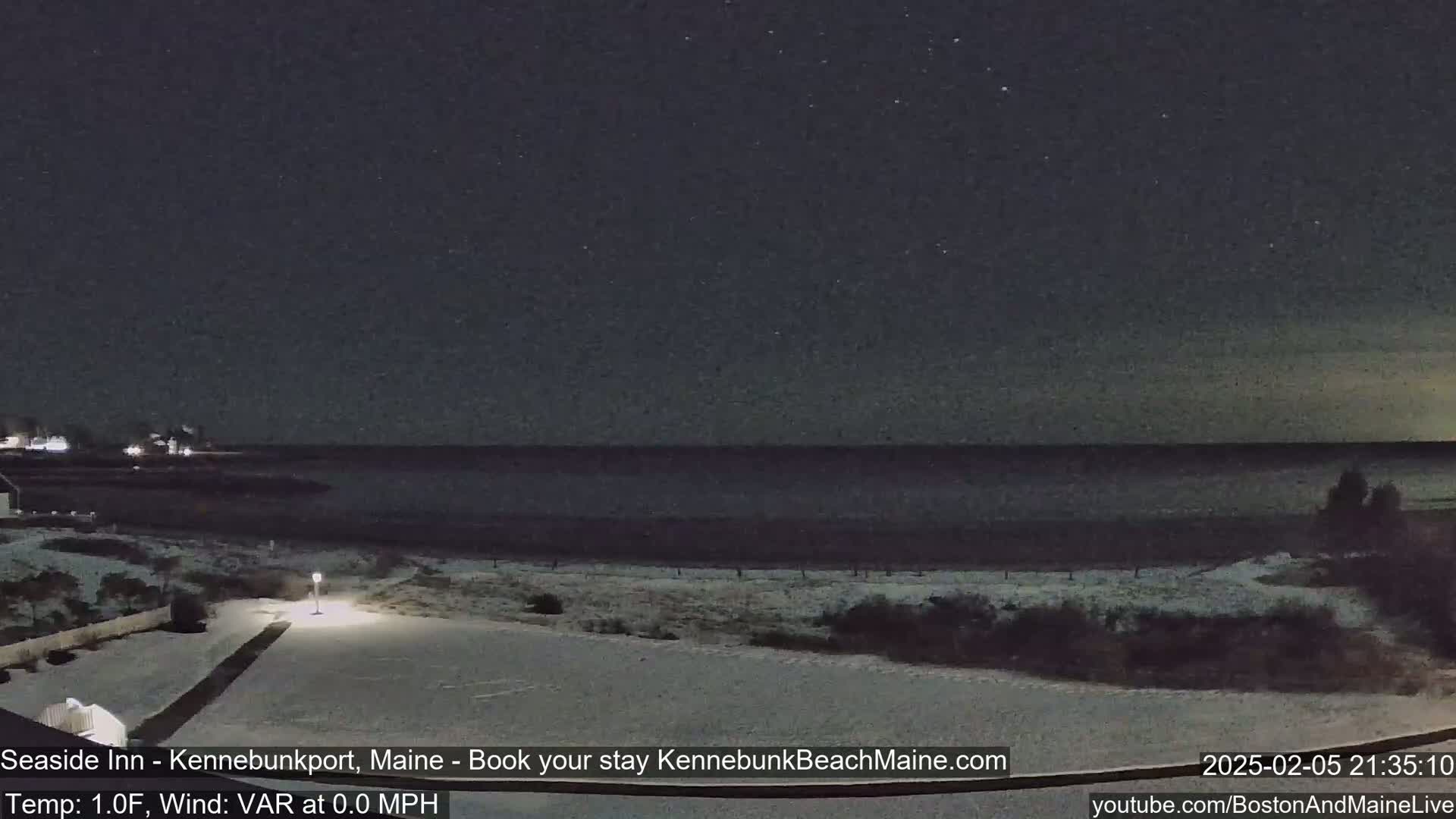 A snow-covered landscape at night shows a dark ocean under a mostly clear sky, with a lit pathway leading to a building in the foreground.