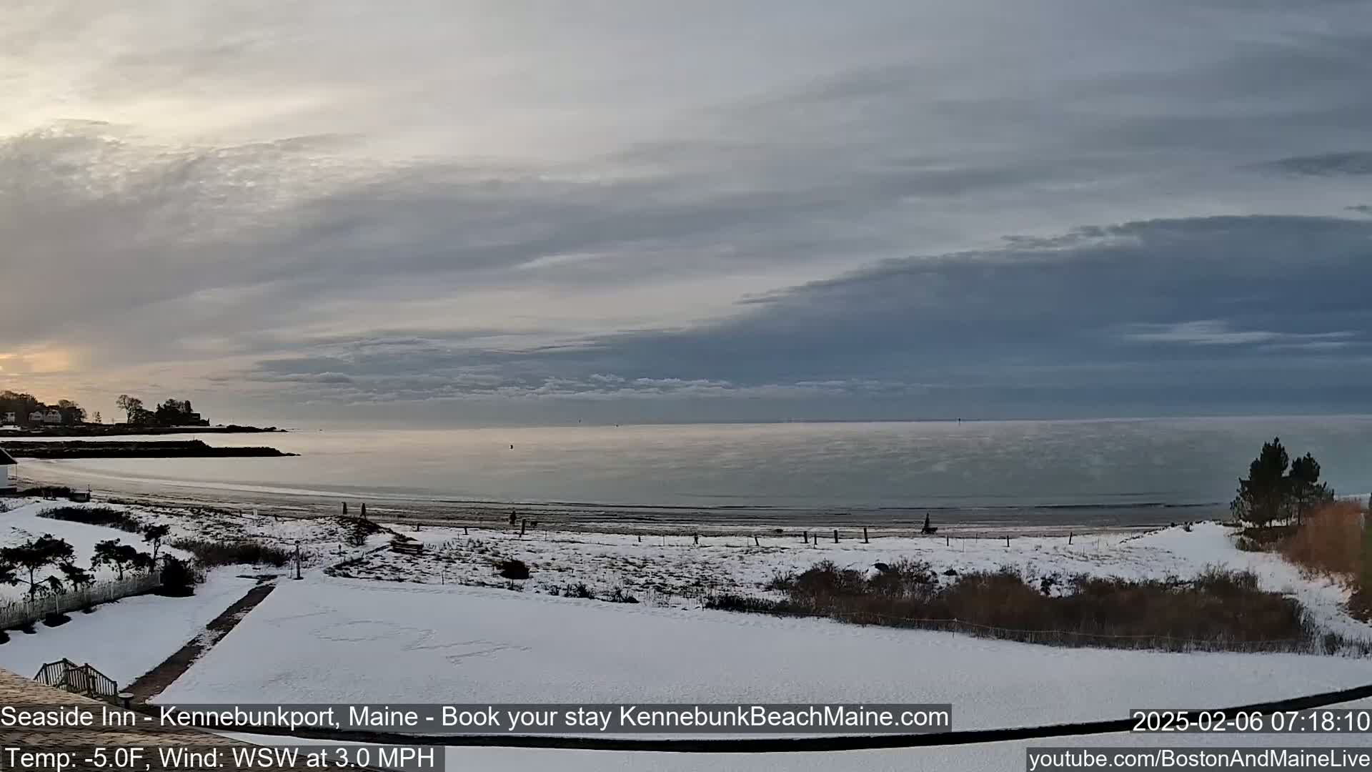 A snow-covered beach and shoreline are visible under a mostly cloudy sky, with a calm ocean in the background.