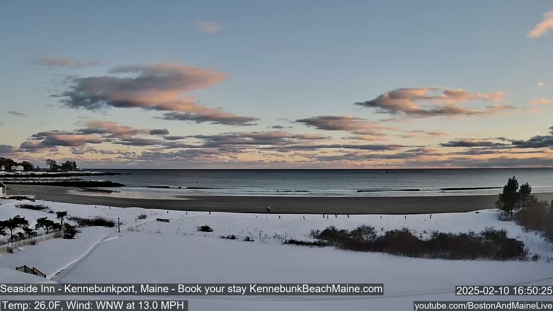 A snow-covered beach scene shows a calm ocean under a partly cloudy, pale pink and blue sky.