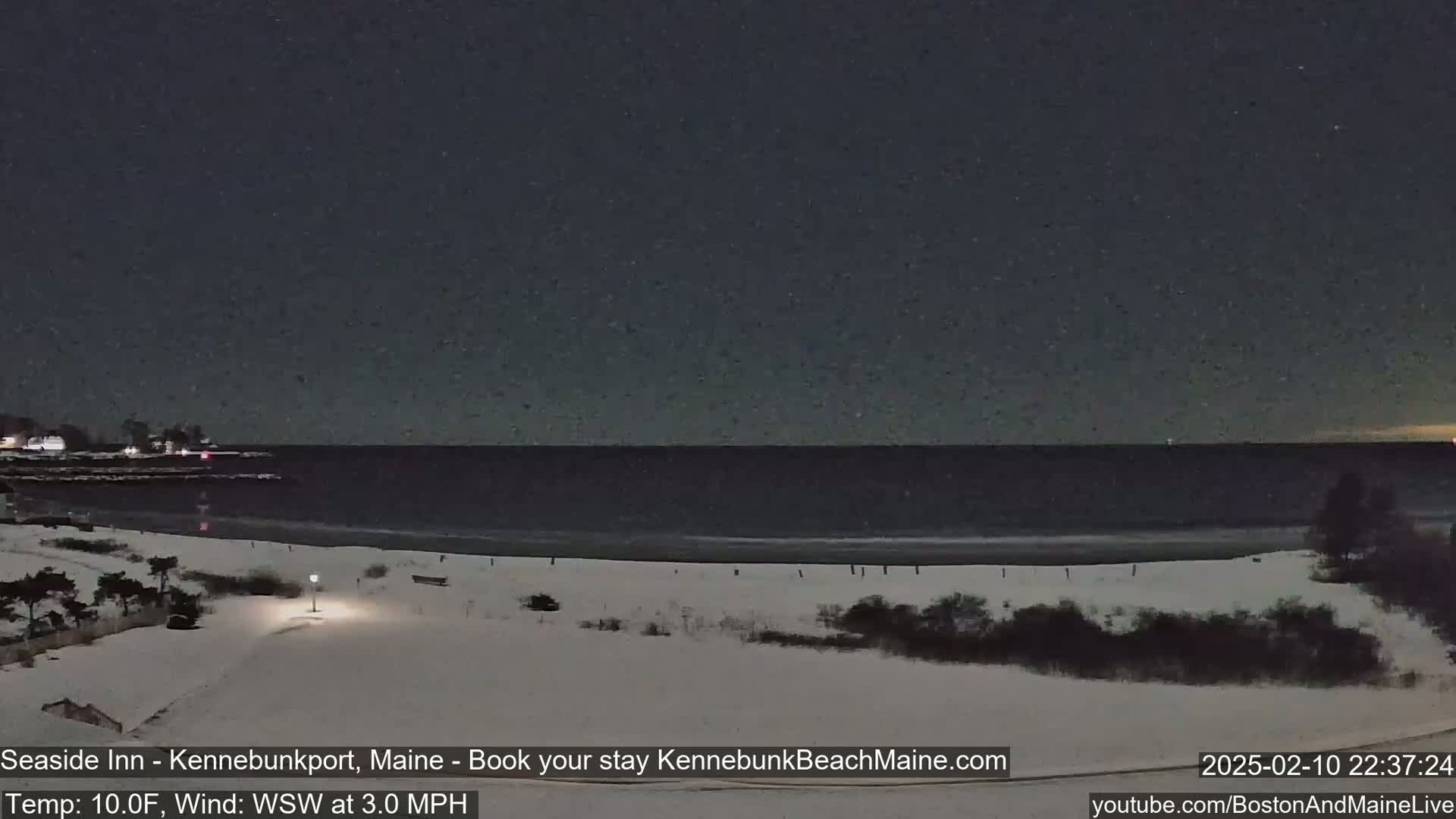 A snow-covered beach at night under a clear, starry sky, with a calm dark ocean in the background and a slight wind.
