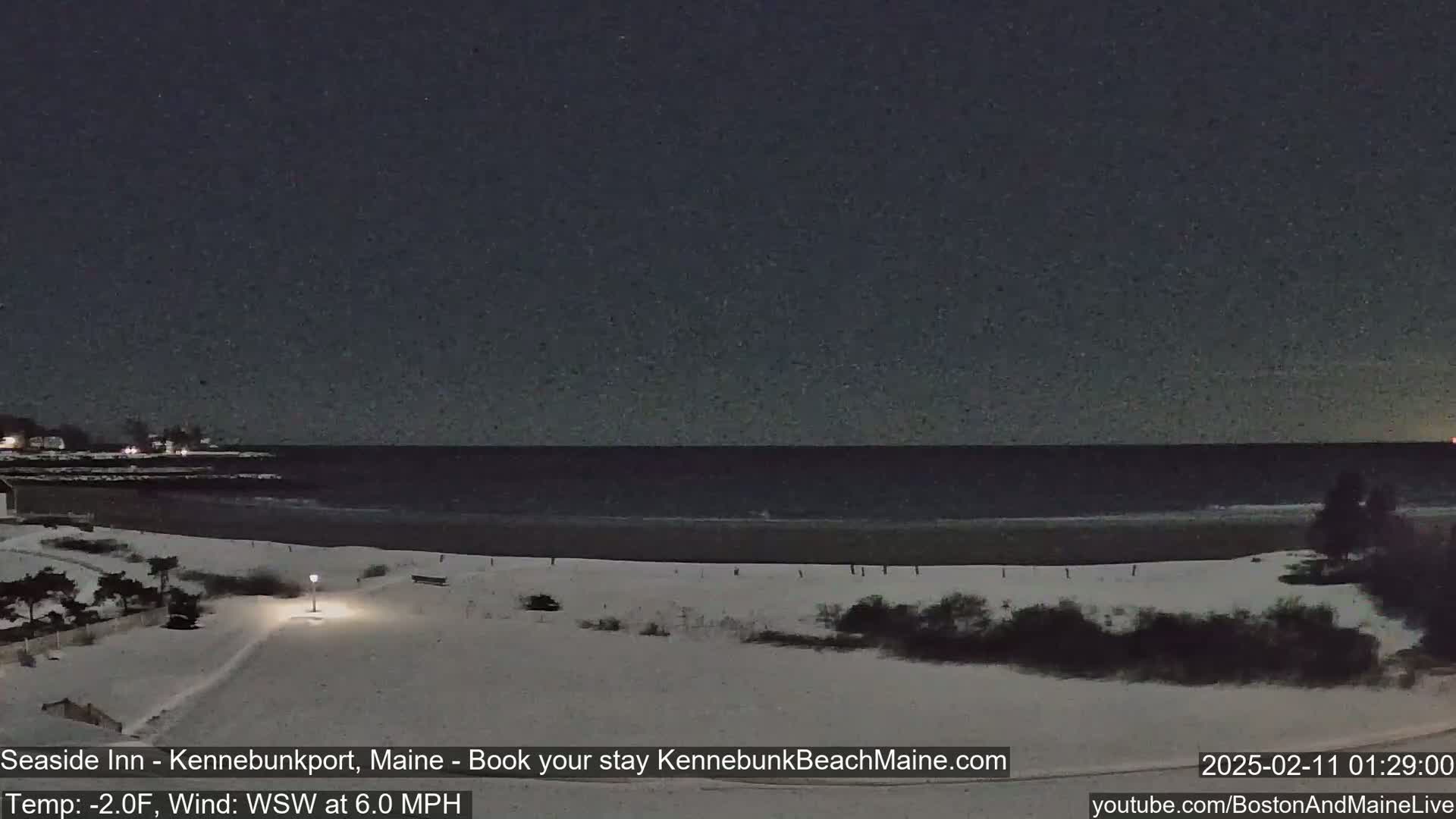 A snow-covered beach at night, with a dark, calm ocean and a few lights visible in the distance under a clear, starry sky.