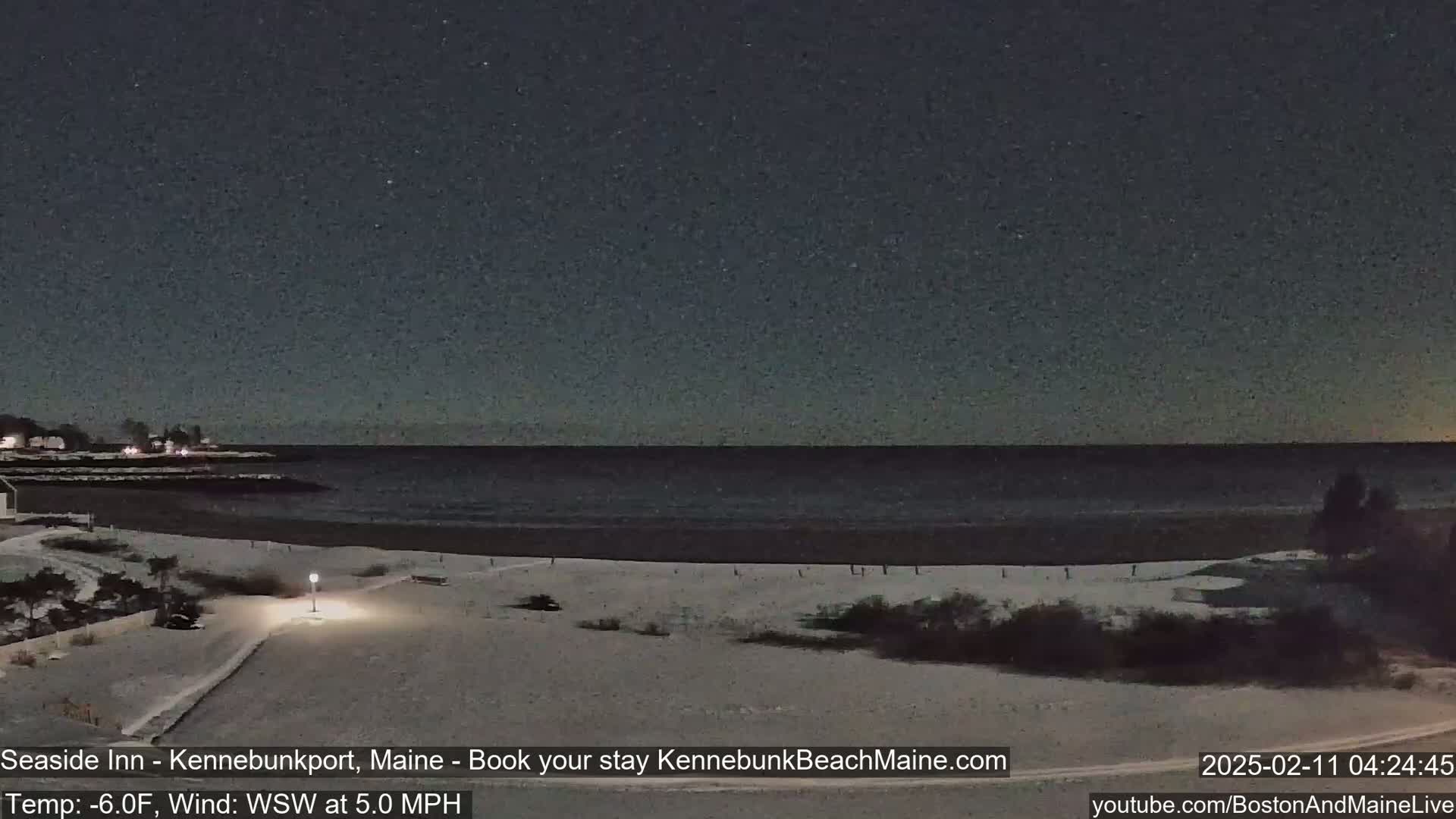 A snow-covered beach at night under a dark, starry sky, with a calm ocean in the background and a slight wind.
