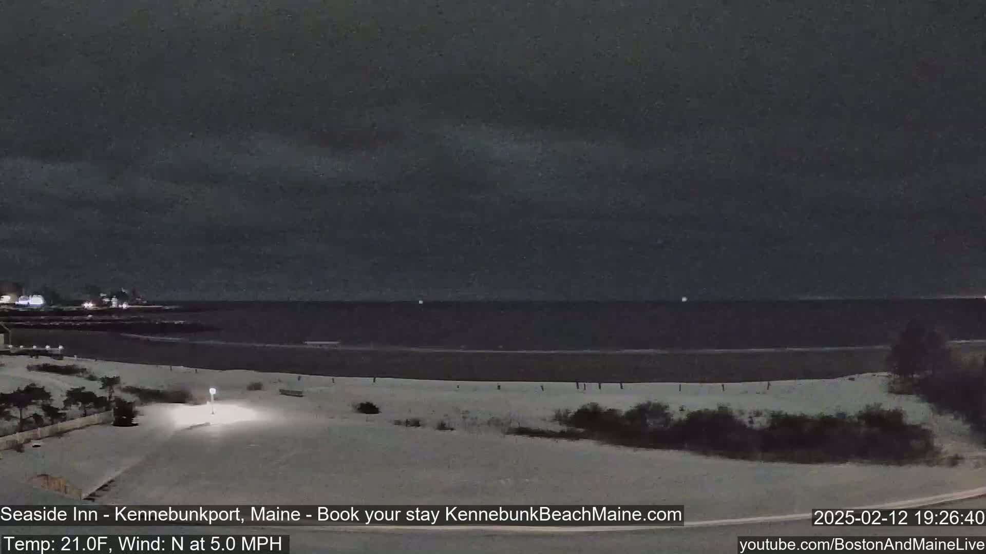 A snow-covered beach at night is seen under a dark, cloudy sky with a calm ocean in the background.