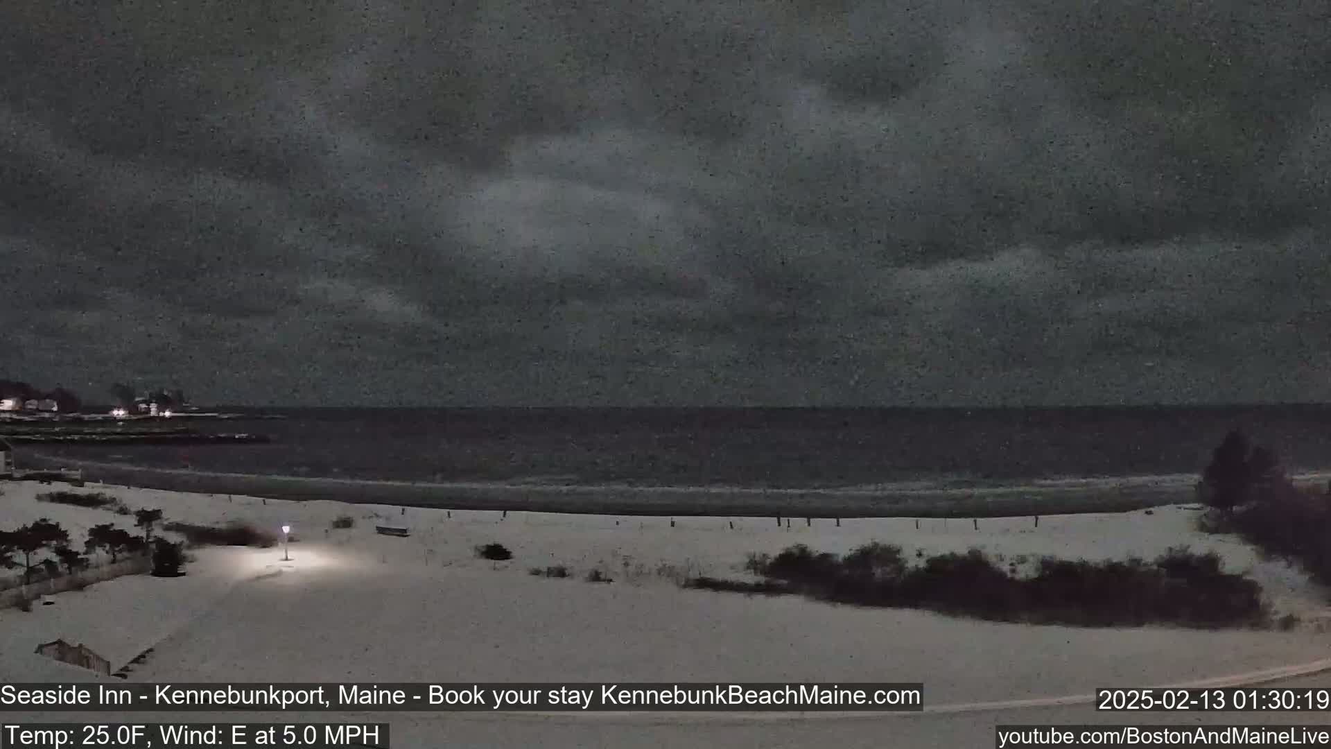 A snow-covered beach at night is seen under a dark, cloudy sky, with a calm ocean in the background.