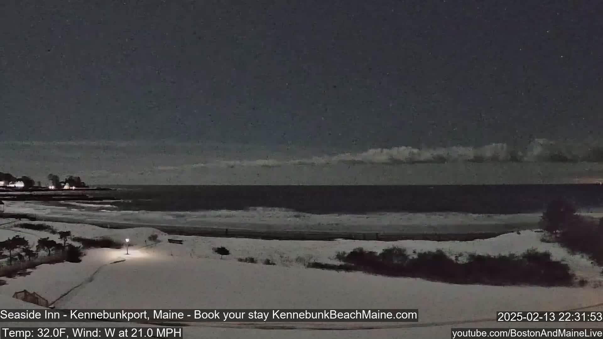 A snow-covered beach at night, with a dark ocean showing small waves under a partly cloudy sky, and a light dusting of snow falling.