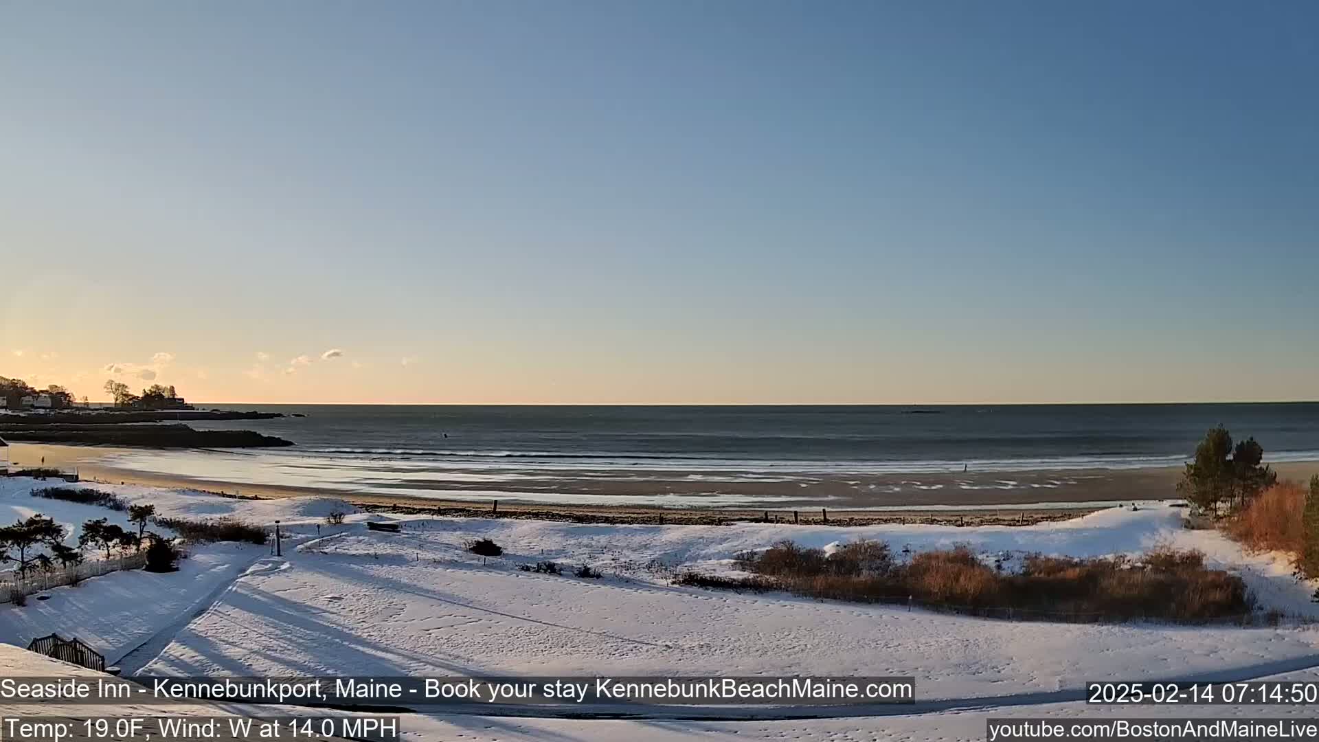 A snow-covered beach with a calm ocean under a clear, light blue sky at sunrise.