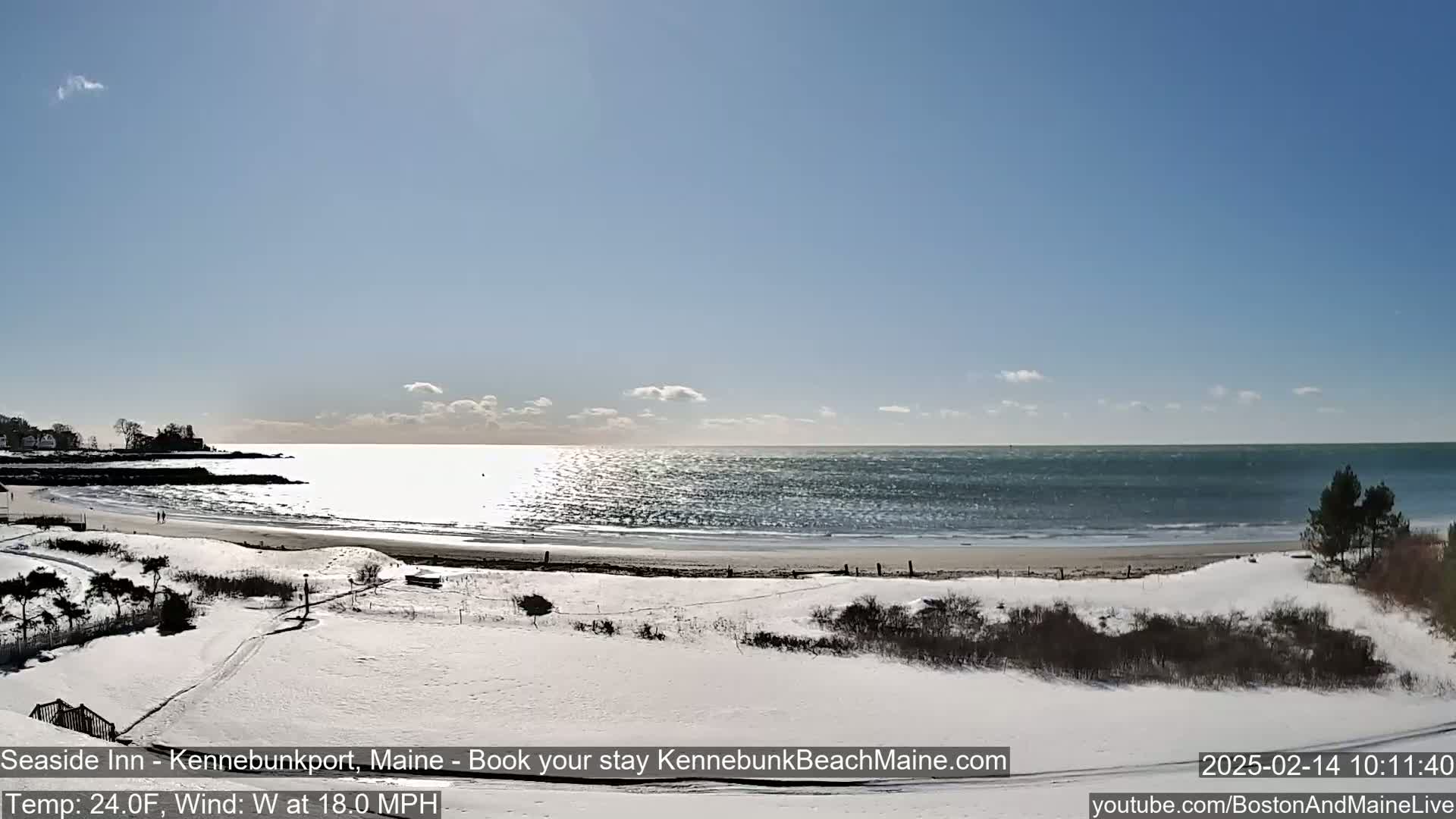 A snow-covered beach scene shows a calm ocean under a mostly sunny sky with a few clouds,  and a slight wind.