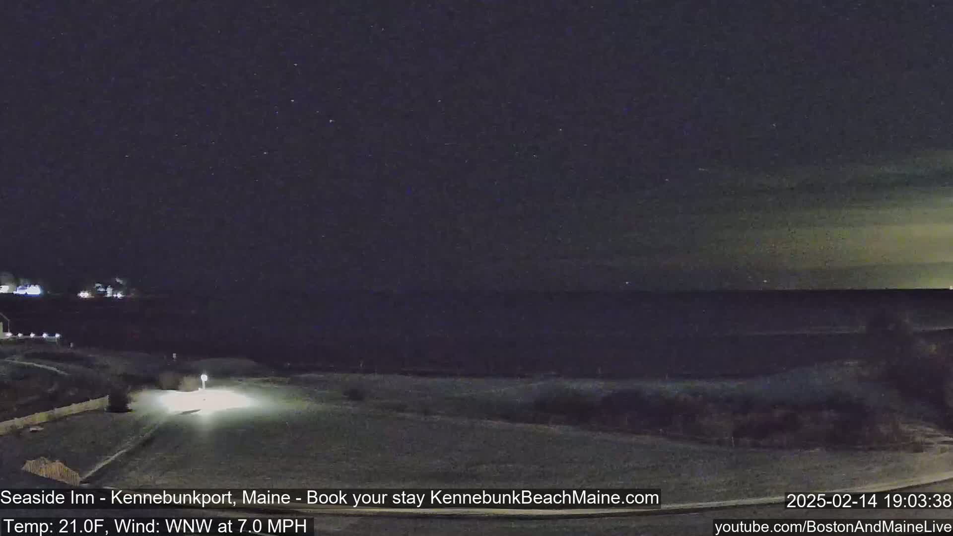 A nighttime view of a snow-covered coastal area shows a dark ocean under a slightly cloudy, starry sky; a road and a vehicle's headlights are visible in the foreground.